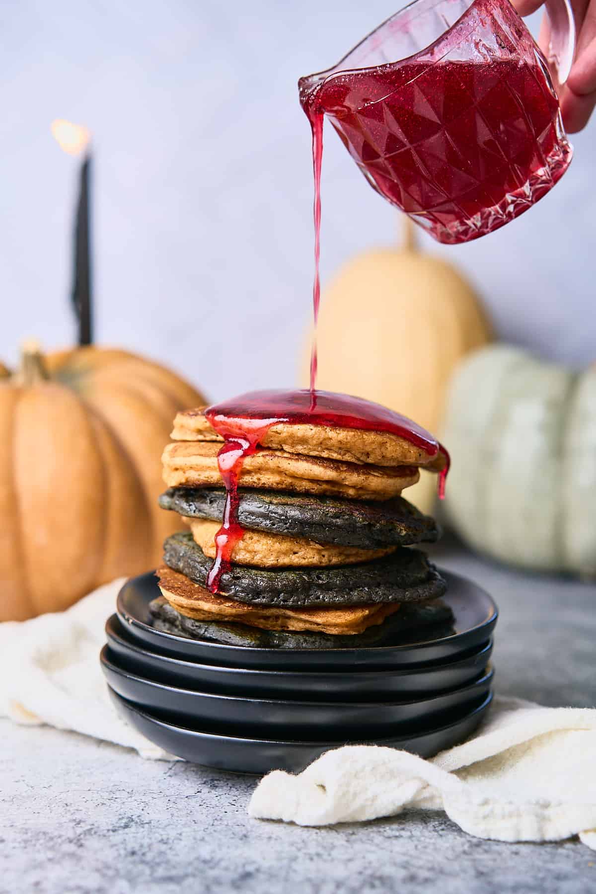 black and orange Halloween pancakes stacked up tall on a stack of black plates, with raspberry bloody syrup being poured onto them, with heirloom pumpkins in the background. 