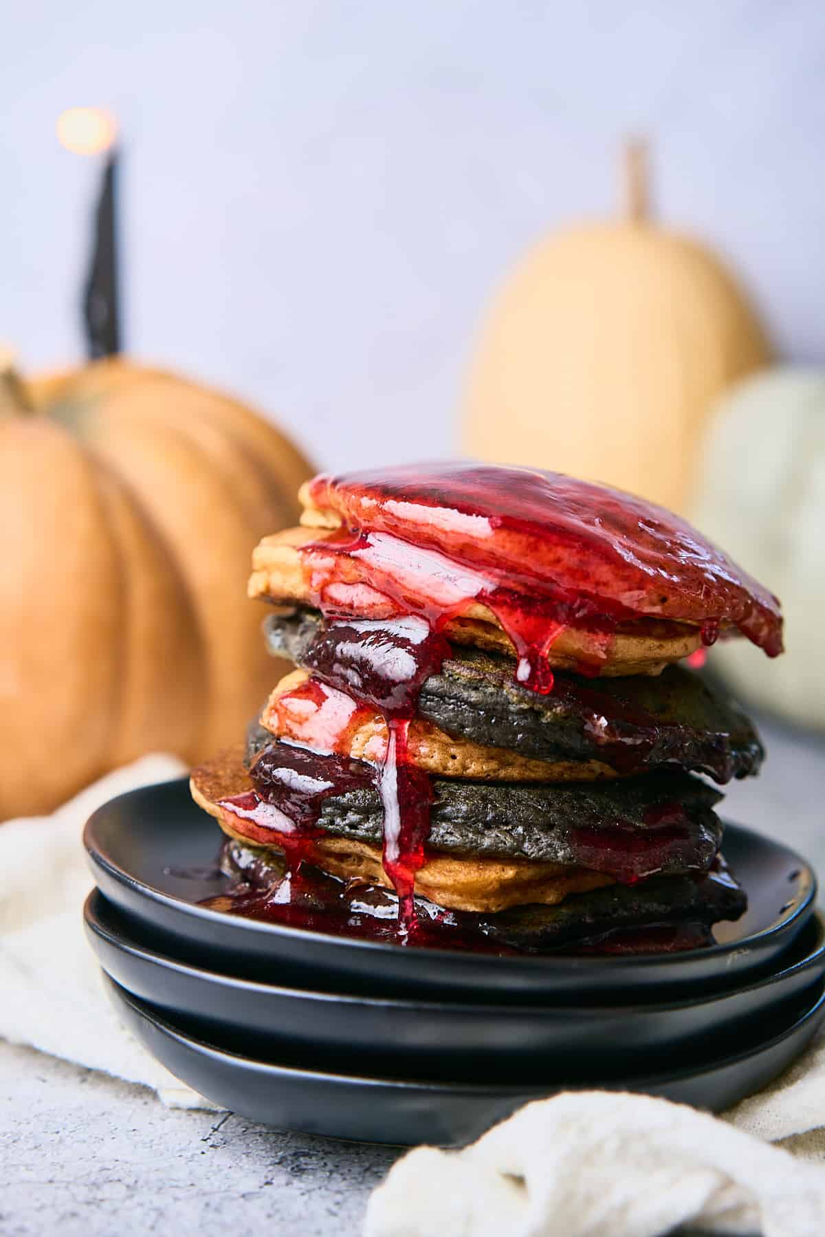 stack of orange and black halloween pancakes dripping with red raspberry syrup to mimic blood on a stack of black plates with heirloom pumpkins in the background. 
