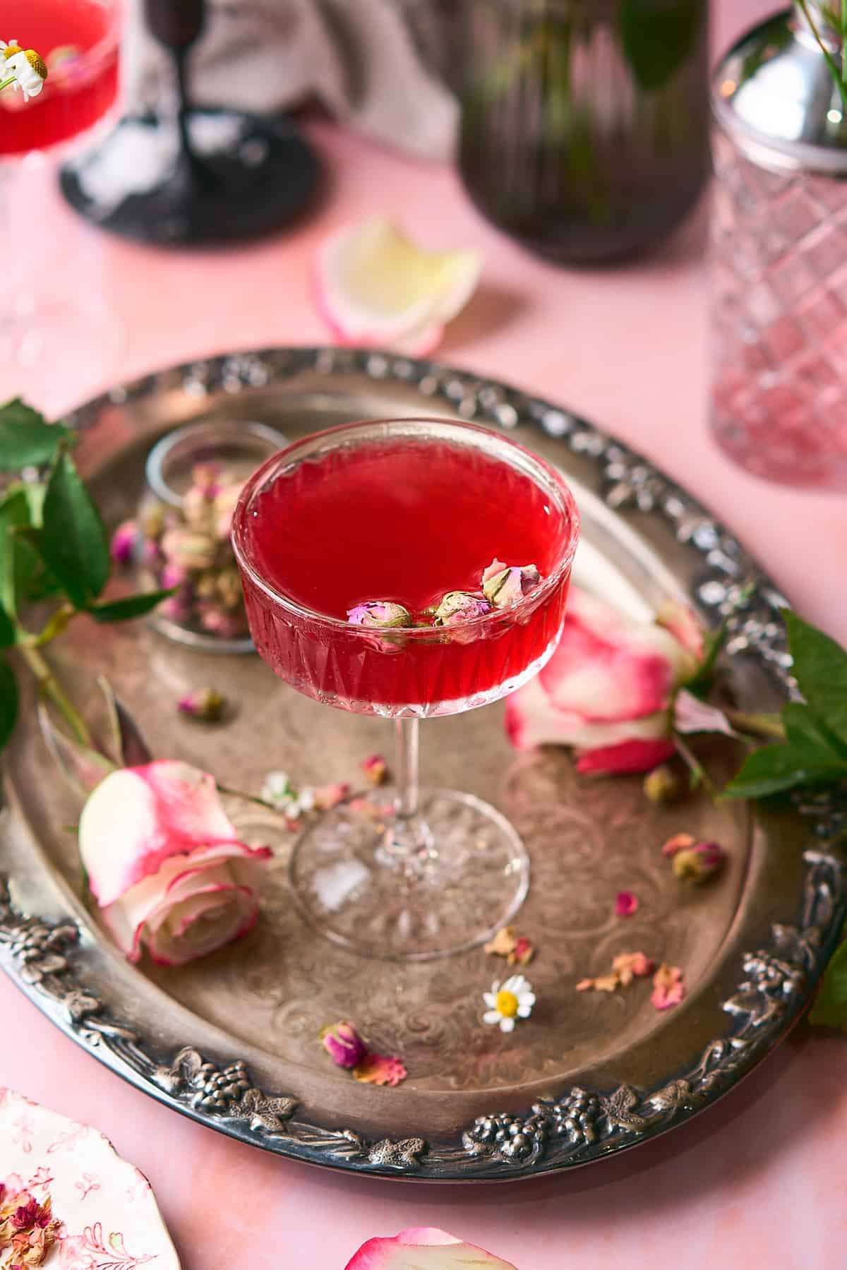 beautiful bright pink cocktail on a silver platter surrounded by roses, with edible rosebuds floating in the drink. 