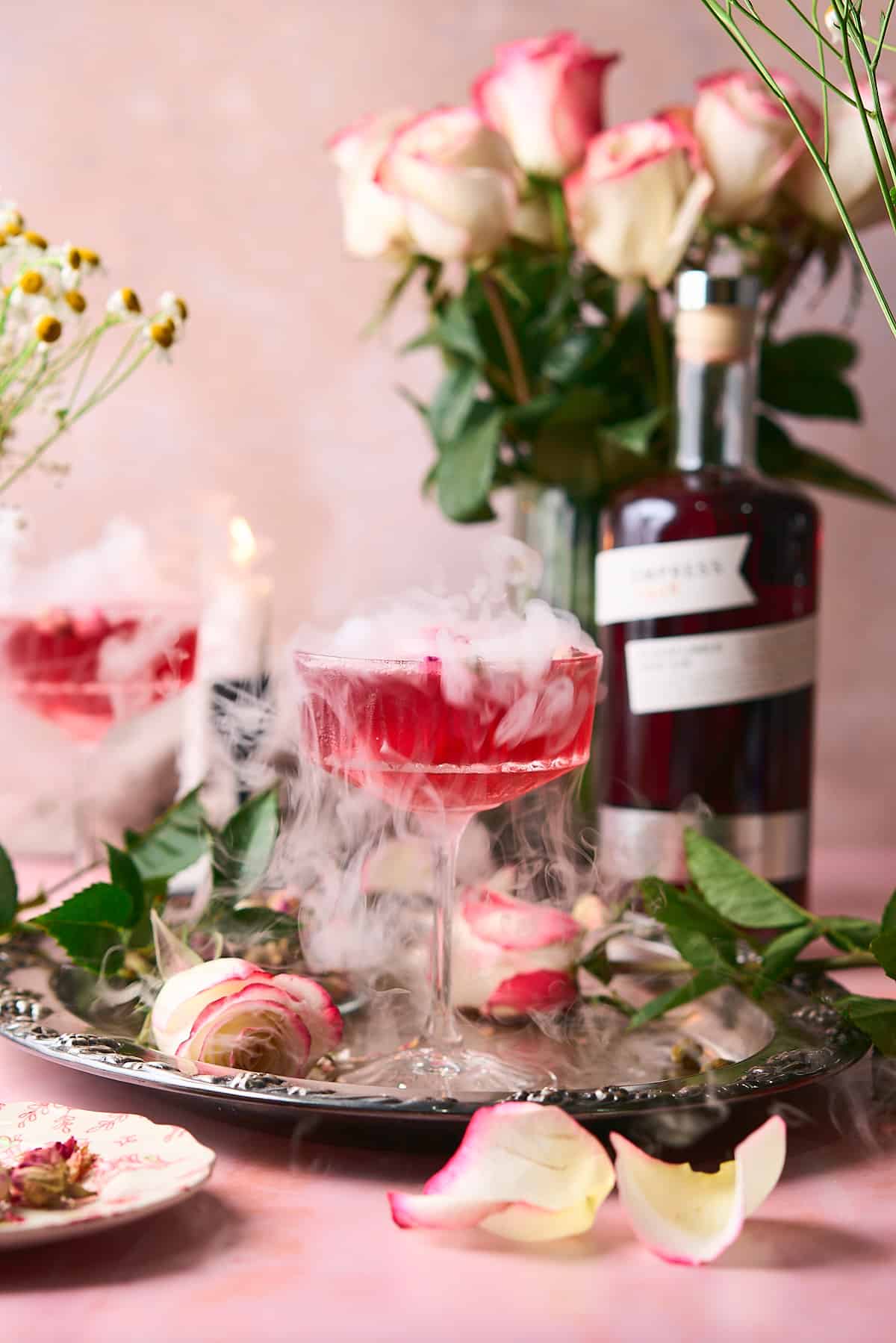 stunning smoking pink cocktail with a candlestick in the background, a bottle of empress pink gin, and roses and fresh flowers surrounding it. 