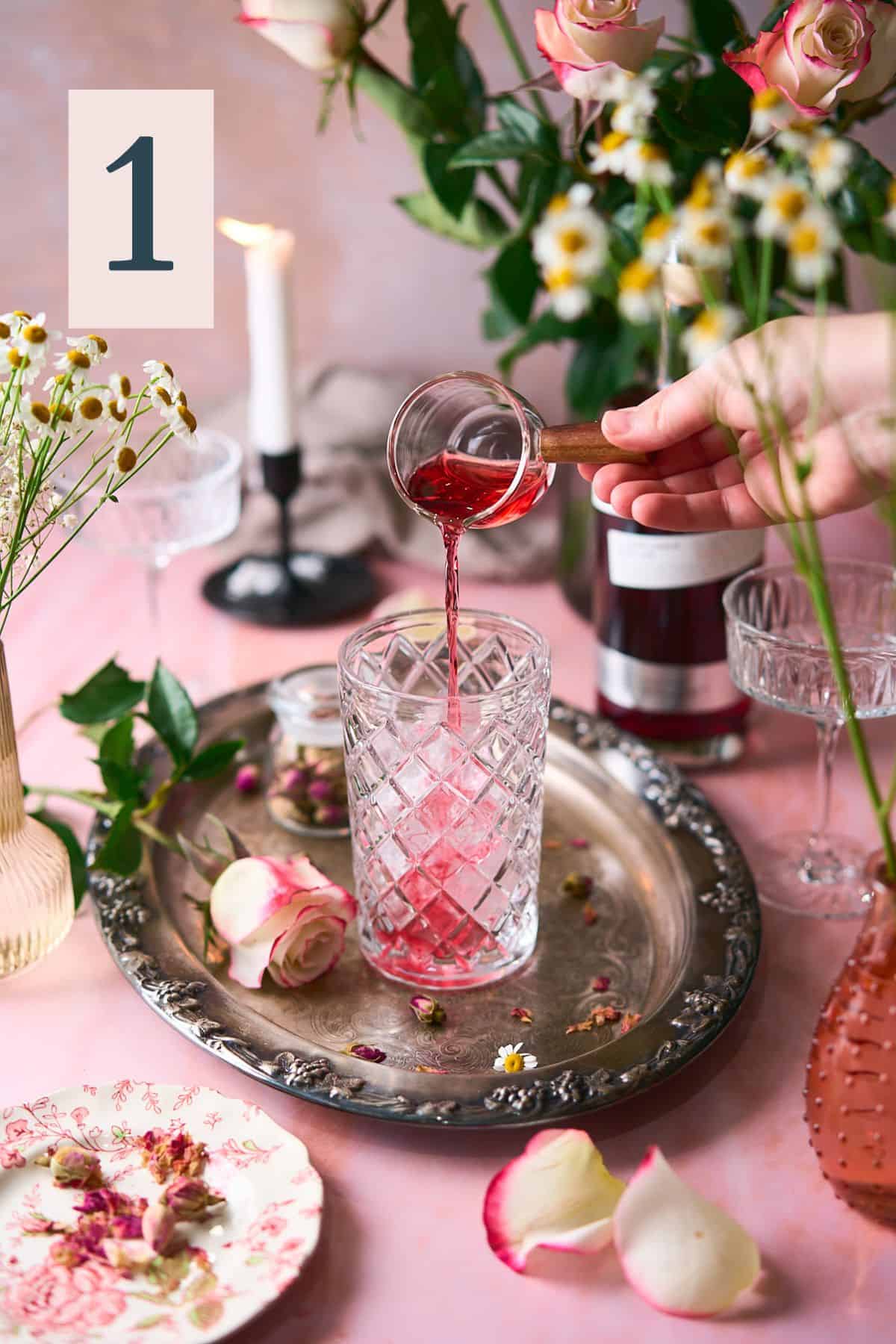 hand pouring cranberry juice into a beautiful glass cocktail shaker with an intricate romantic scene of fresh roses, candles and flowers surrounding it. 