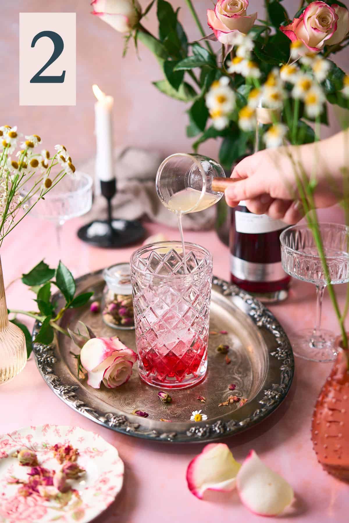 hand pouring lemon juice into a beautiful glass cocktail shaker with an intricate romantic scene of fresh roses, candles and flowers surrounding it.