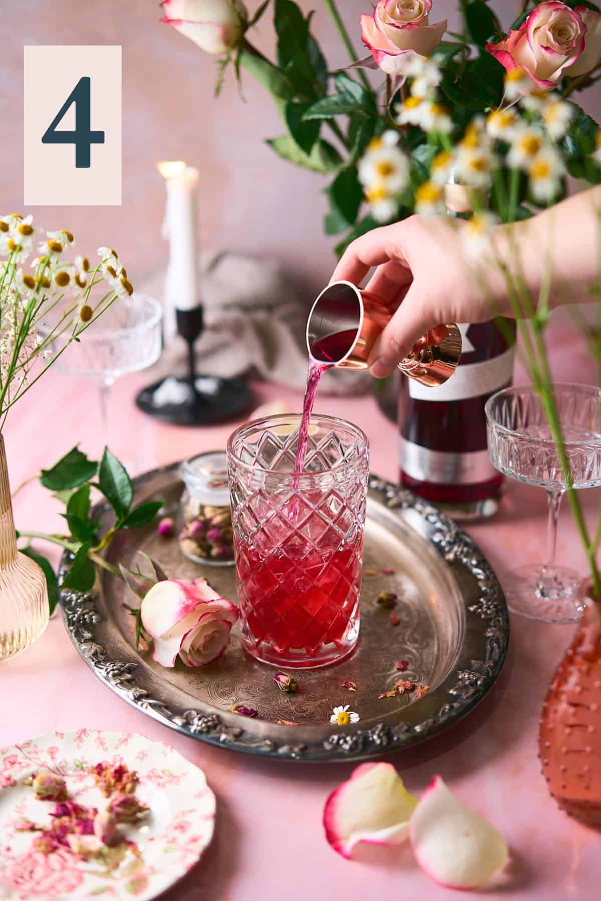hand pouring pink gin into a beautiful glass cocktail shaker with an intricate romantic scene of fresh roses, candles and flowers surrounding it.