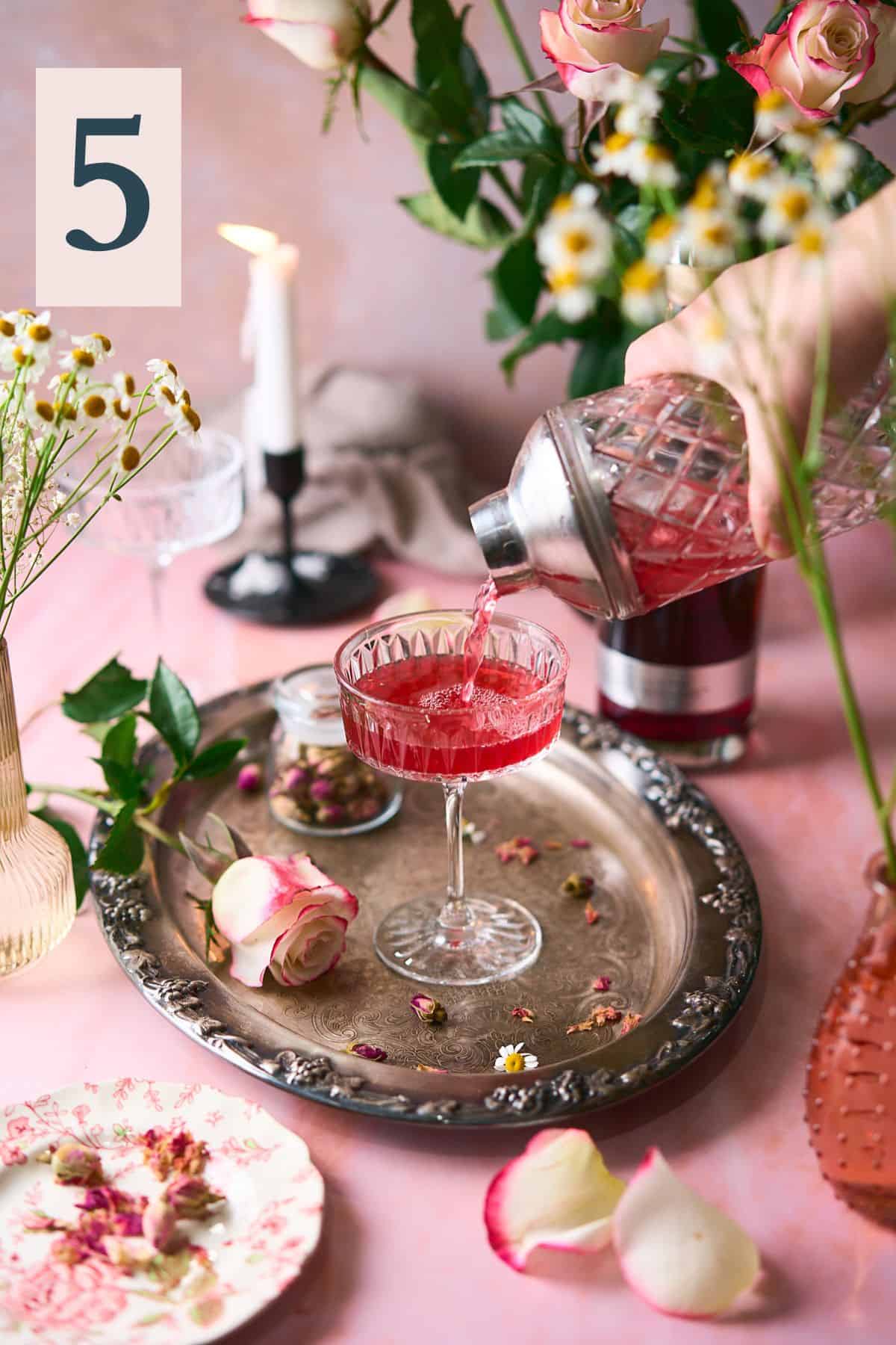 hand pouring a bright pink cocktail from a shaker into a coupe glass with an intricate romantic scene of fresh roses, candles and flowers surrounding it.