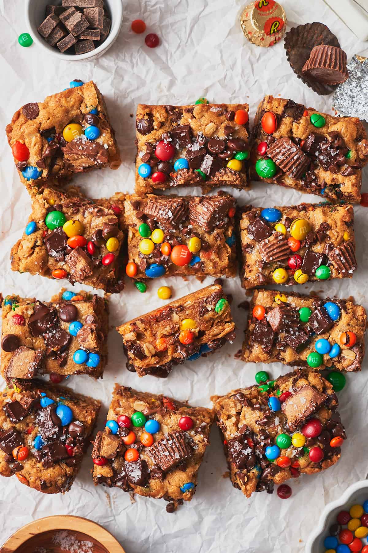 Overhead view of 12 monster cookie bars with M&ms, sea salt, and reese's peanut butter cups on crinkled white parchment paper. 