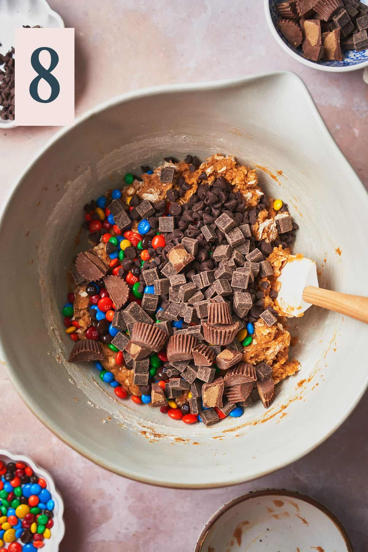 M&ms, chocolate chips, chunks, and reese's peanut butter cups in a bowl of cookie dough batter. 