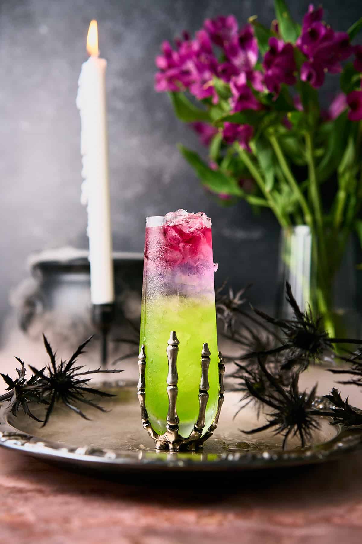 Colorful layered green, purple and pink cocktail in a skeleton glass with a witches cauldron leaking dry ice in the background. 
