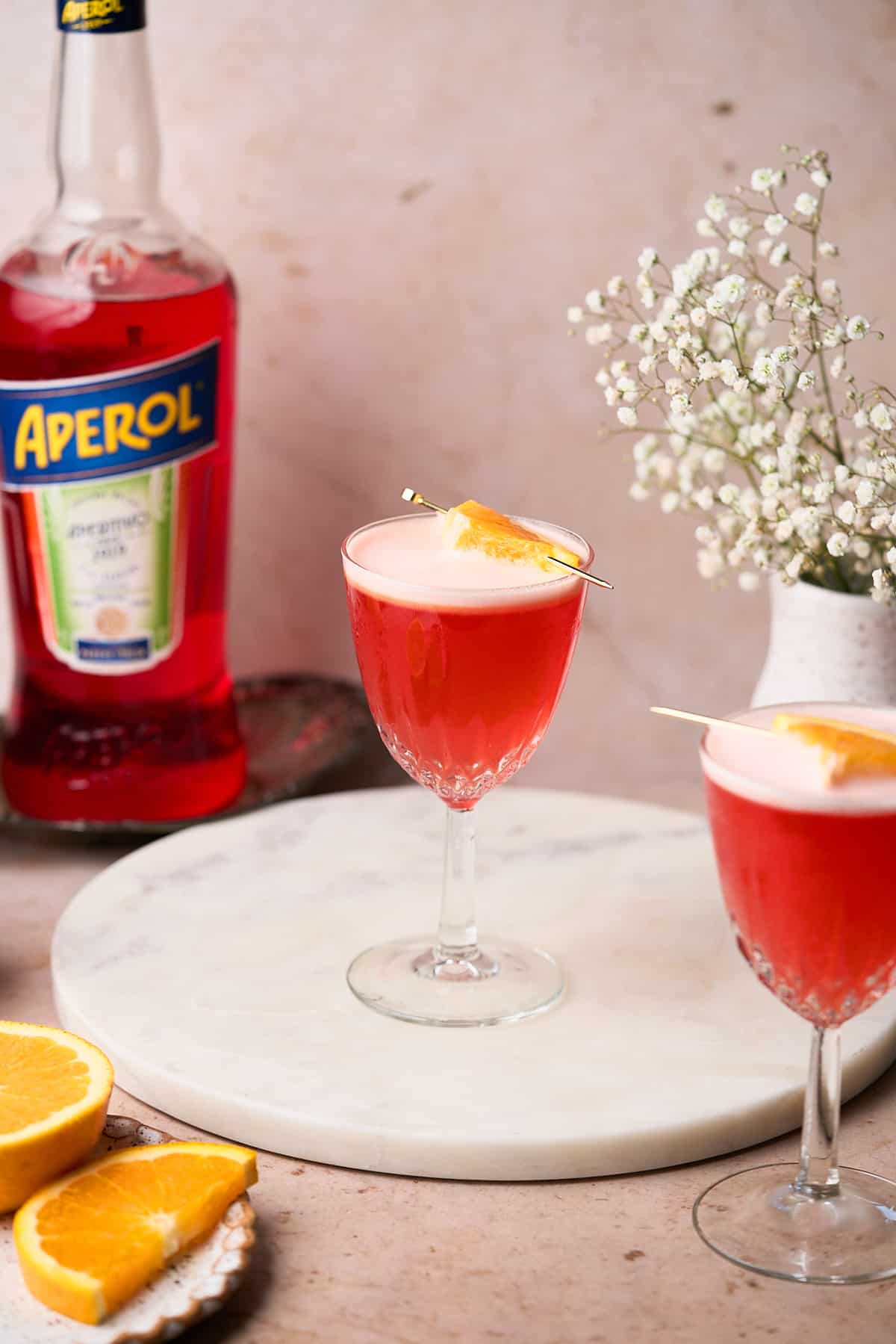 two aperol sours on a round white marble serving platter, with a bottle of aperol in the background, with little white baby's breath flower. 