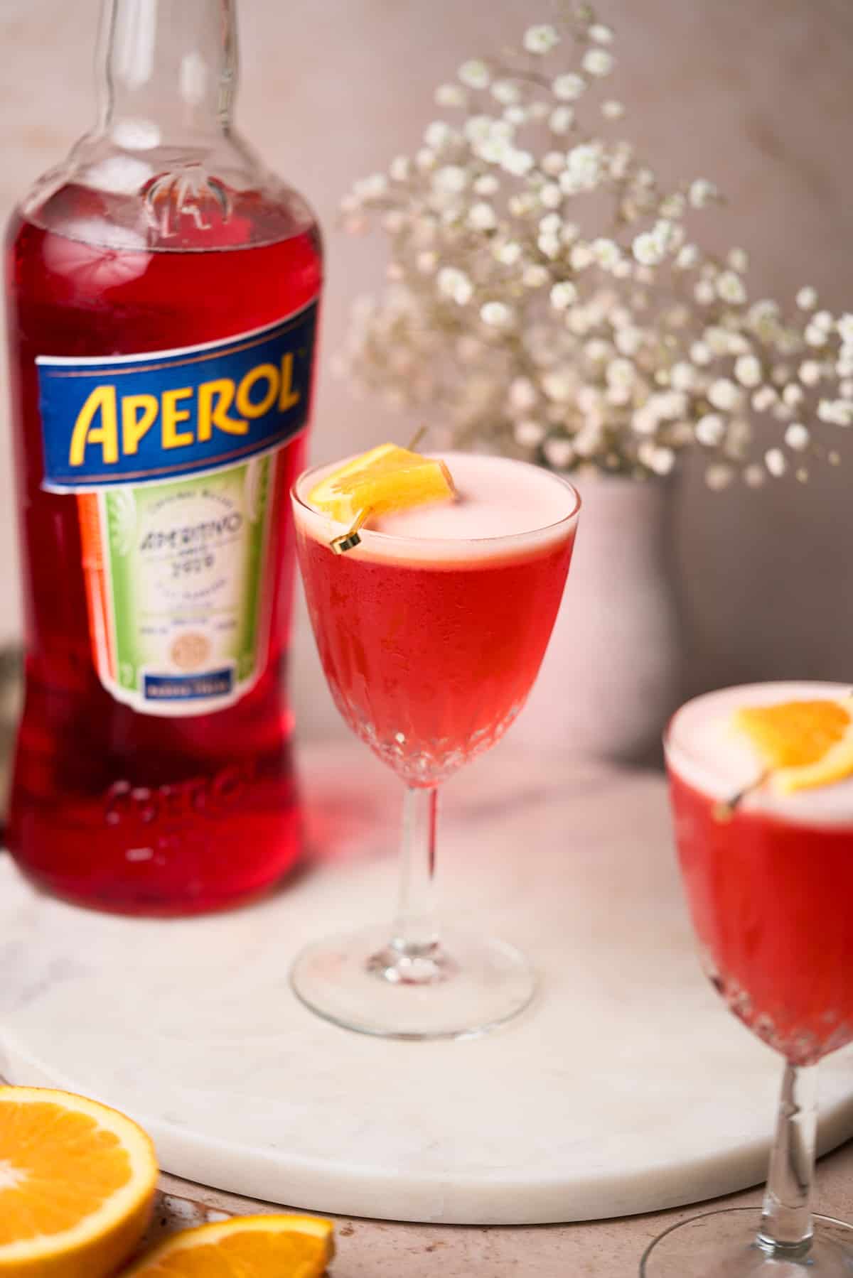 two aperol sour cocktails with a foamy top, and a small wedge of orange on top of the cocktail with a bottle of Aperol and white baby's breath flowers in the background.