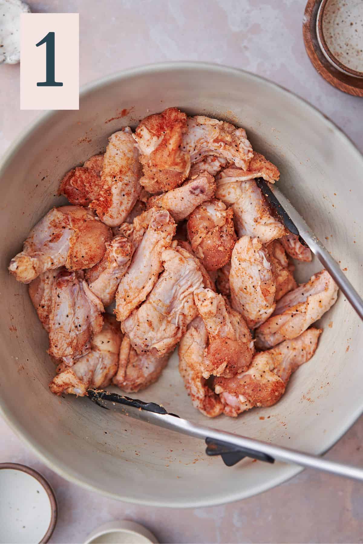 Split chicken wings tossed in seasonings with tongs inside a bowl.
