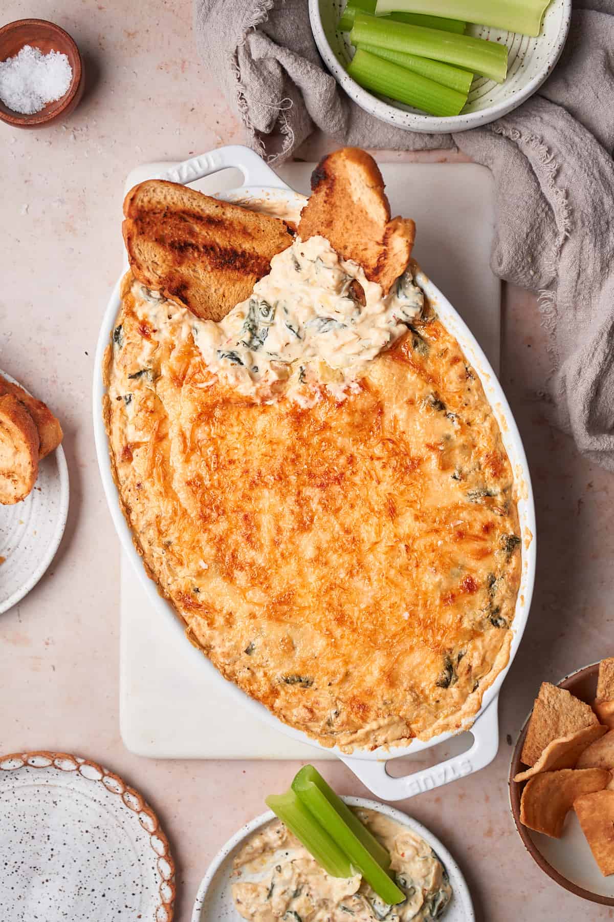 Baked spinach artichoke dip in an oval baking dish surrounded by dipping vessels, with bread in the baking dish to reveal the inside of the dip. 