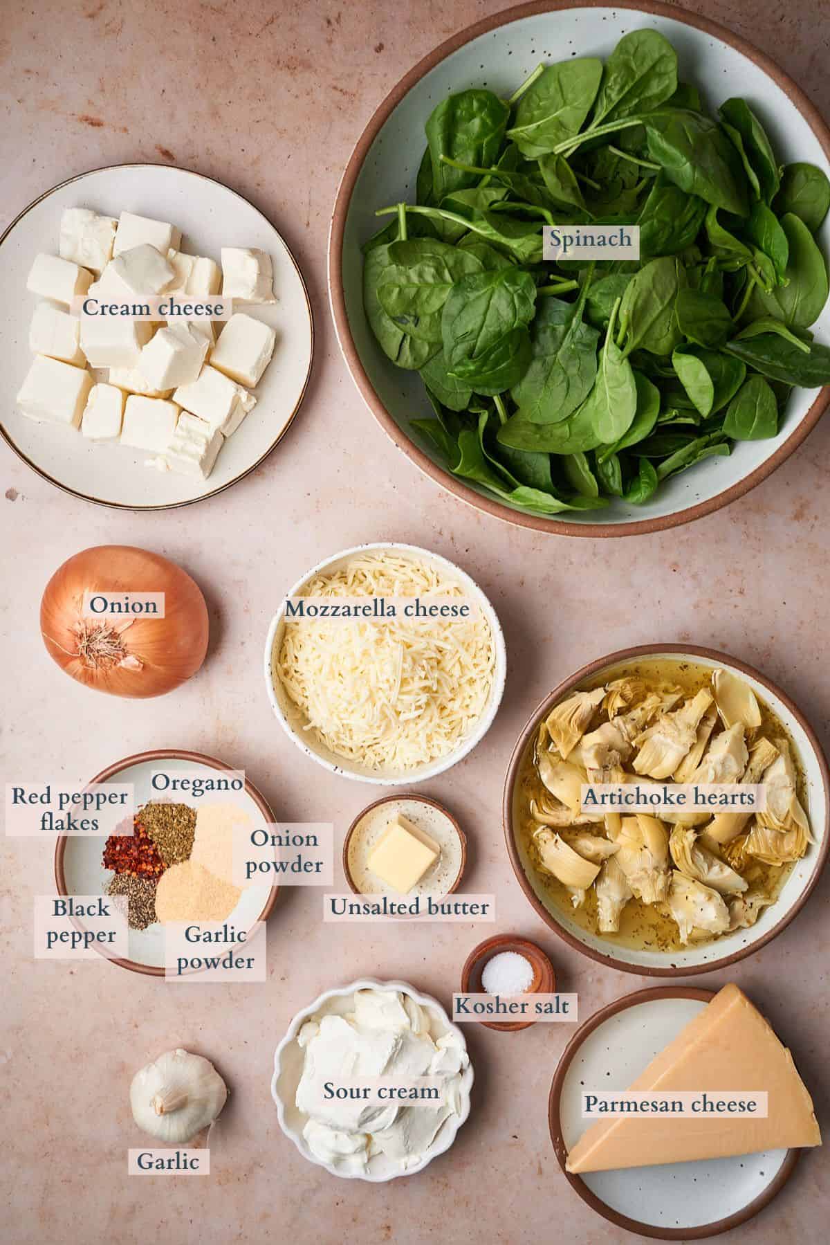 Ingredients to make baked spinach artichoke dip laid out on a table and labeled to denote each ingredient. 