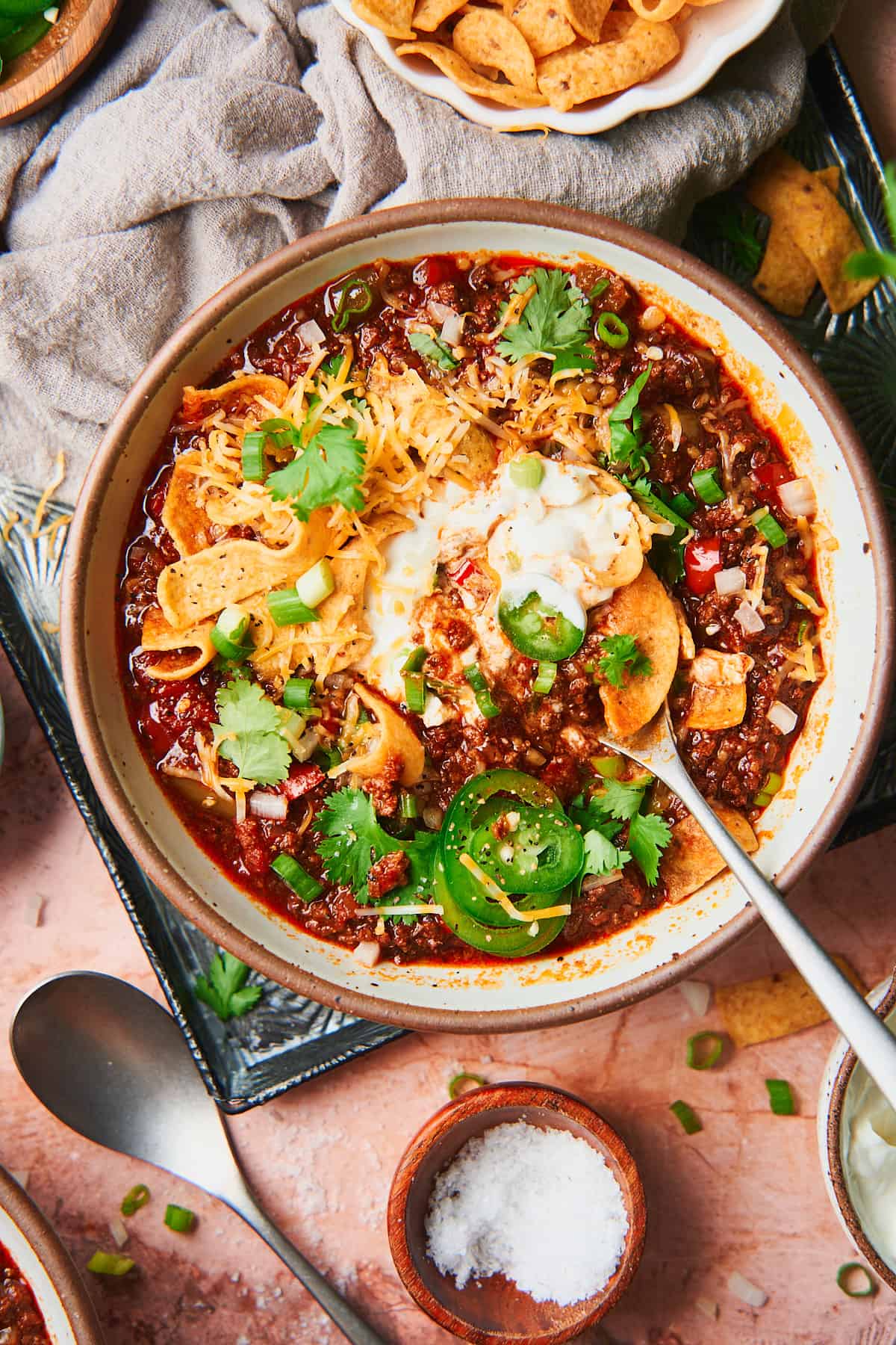 big bowl of beanless chili topped with fritos, sour cream, jalapenos, cheese, and cilantro on a warm textured back ground. 