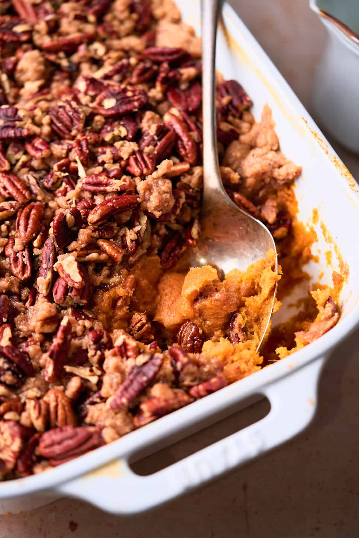 close up shot of sweet potato casserole in a large square baking dish with a spoon, showing the inside of the dish with a pecan topping.