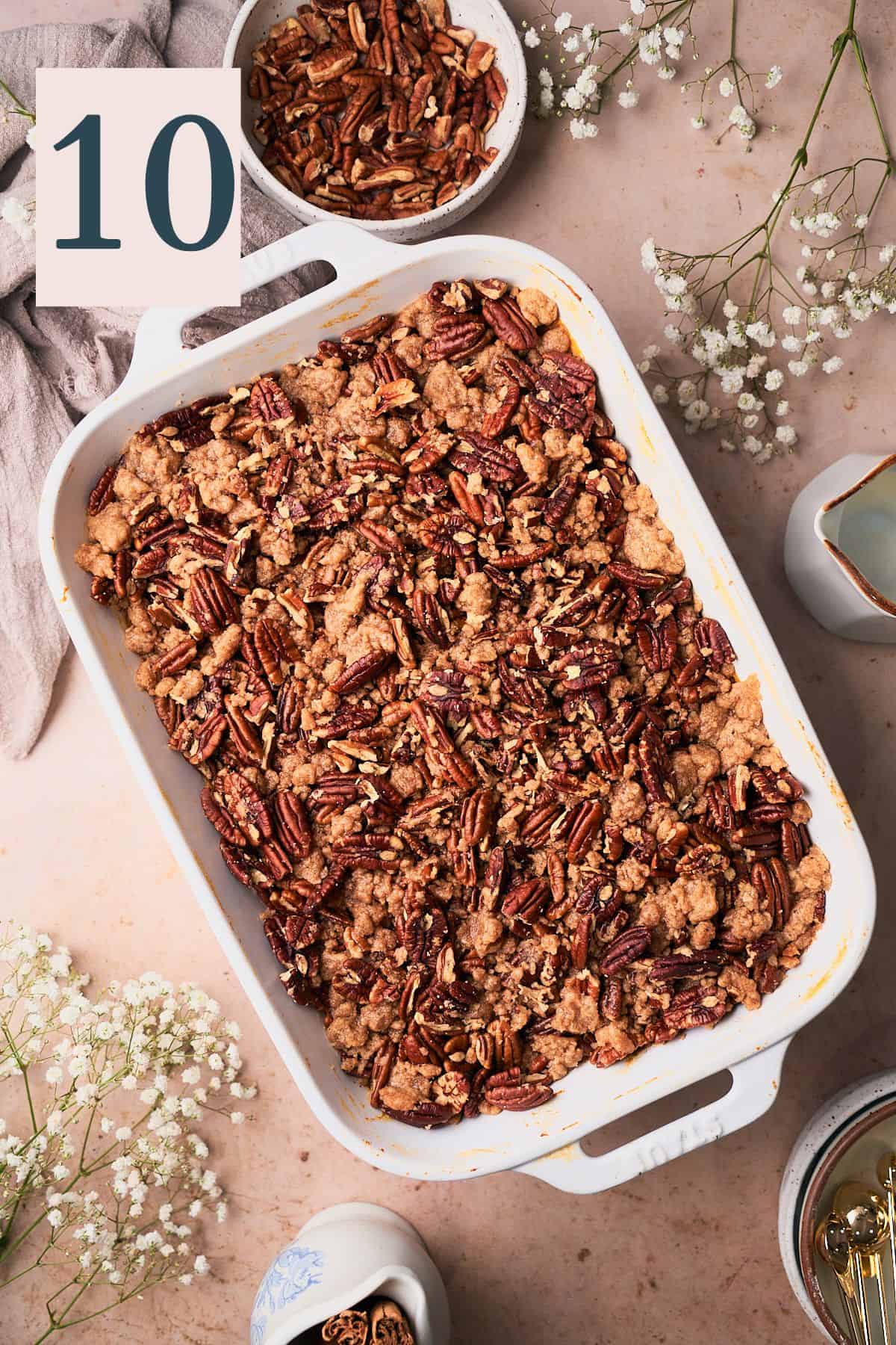 baked sweet potato casserole in a baking dish and ready to serve, surrounded by small white flowers, pecans, and cinnamon sticks.