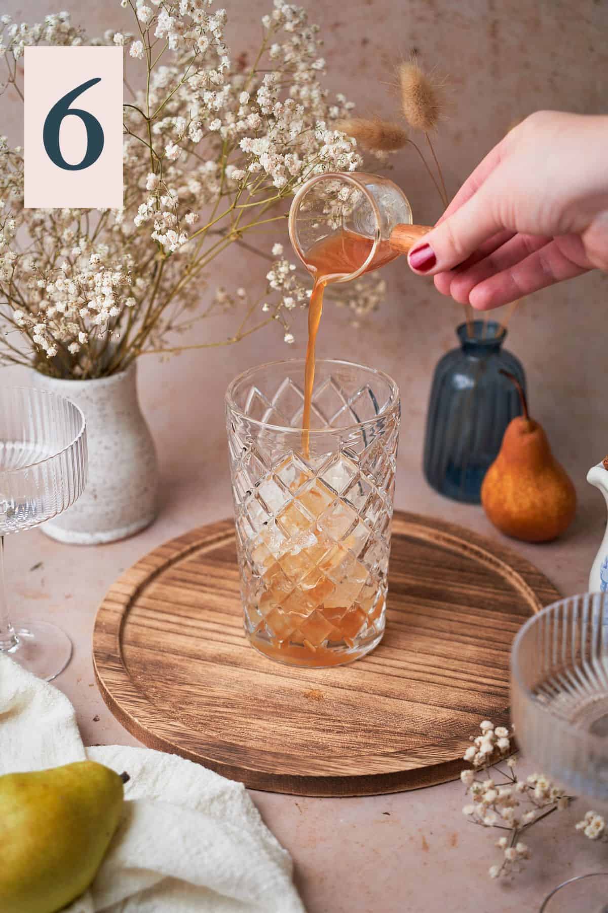 hand pouring spiced simple syrup into a cocktail shaker surrounded by pears, flowers, and a white linen napkin.