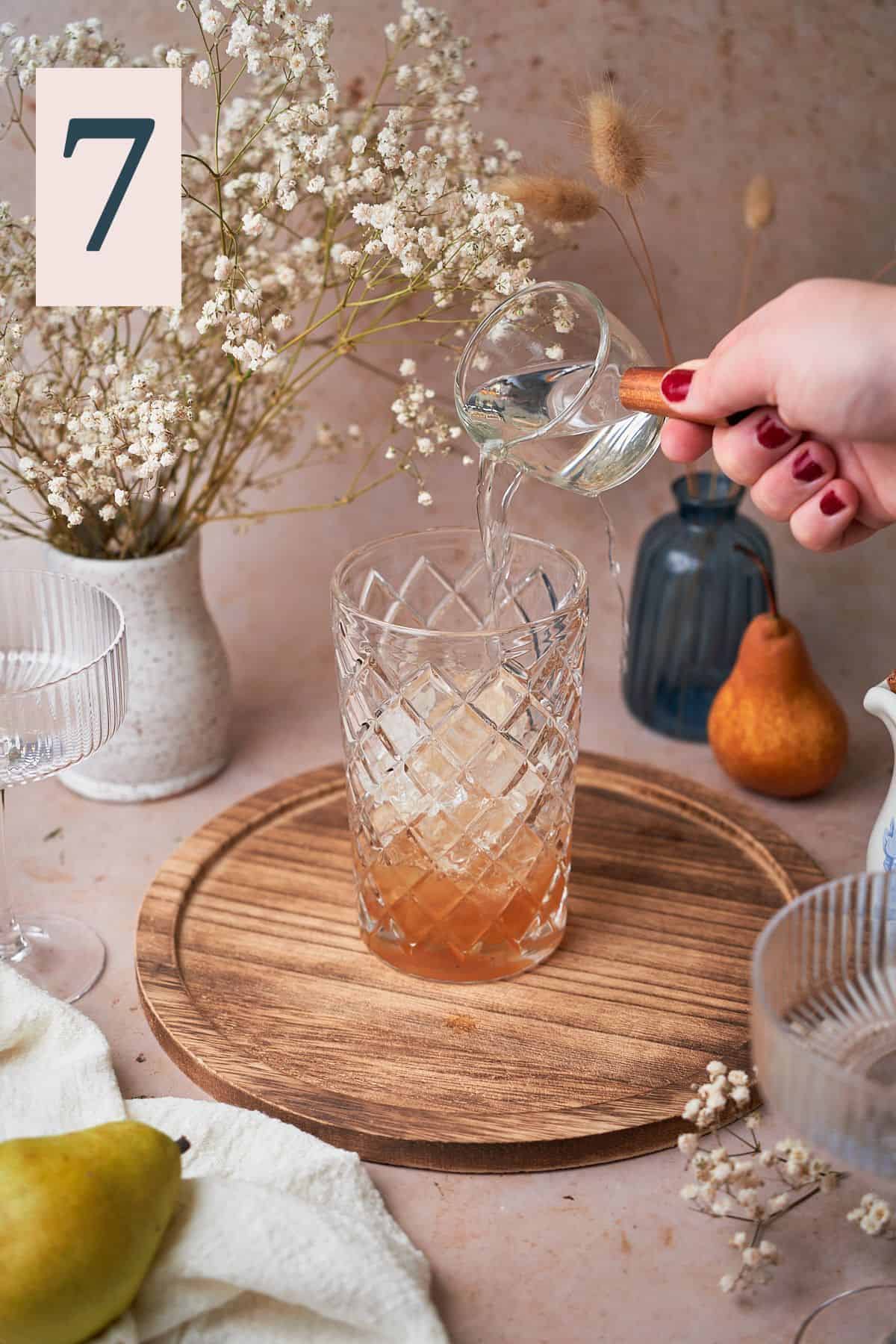 hand pouring vodka into a cocktail shaker surrounded by pears, flowers, and a white linen napkin.