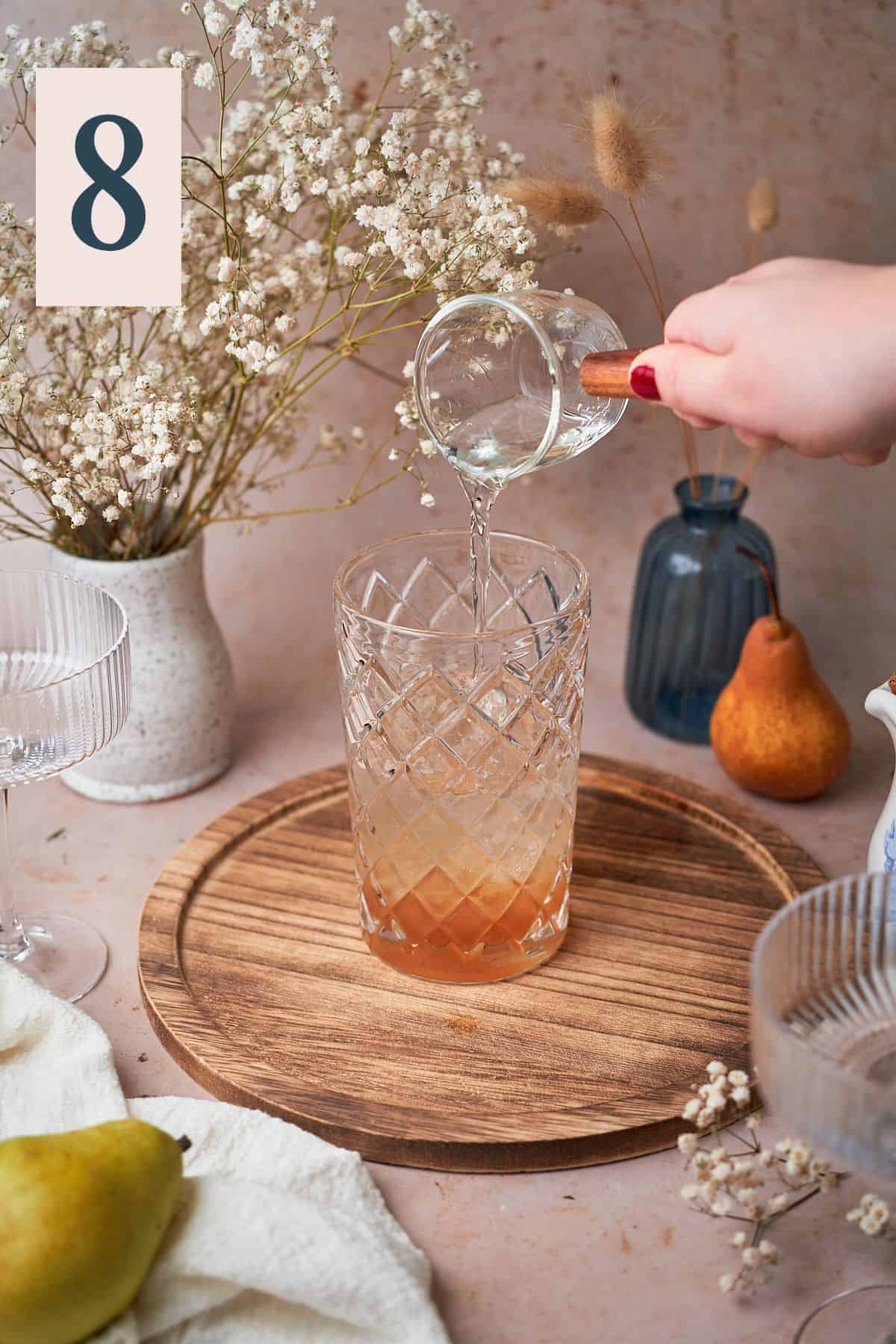 hand pouring orange liqueur into a cocktail shaker surrounded by pears, flowers, and a white linen napkin.