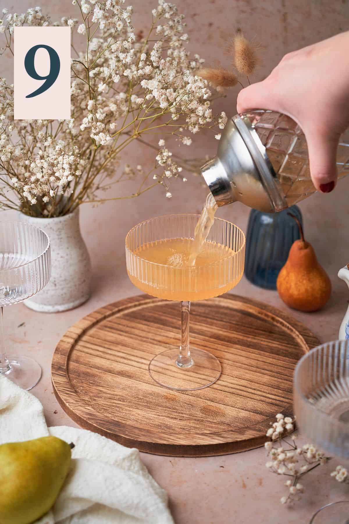 hand pouring a cocktail into a coupe glass into a cocktail shaker surrounded by pears, flowers, and a white linen napkin.