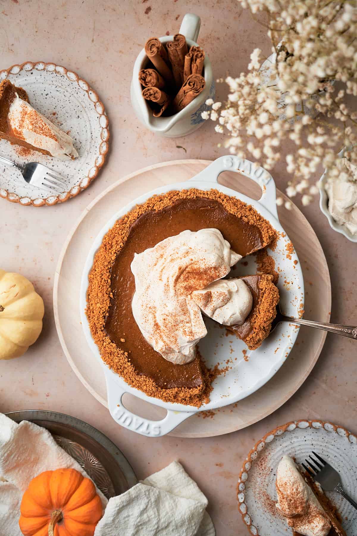 overhead scene of a pumpkin pie topped with a whipped cream topping, with some slices taken out and placed on surrounding plates, with flowers in the corner, small pumpkins, and cinnamon sticks. 