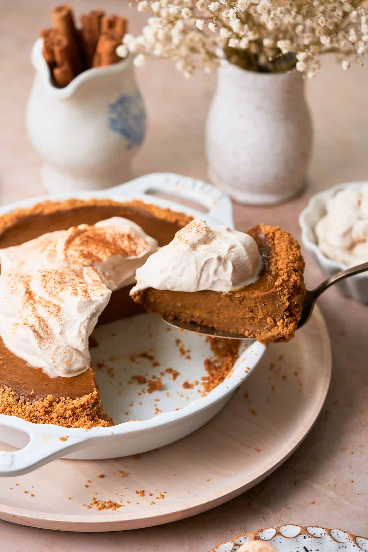 stunning shot of a slice of pumpkin pie with graham cracker crust being pulled out from a pie plate, that is topped with whipped cream. 