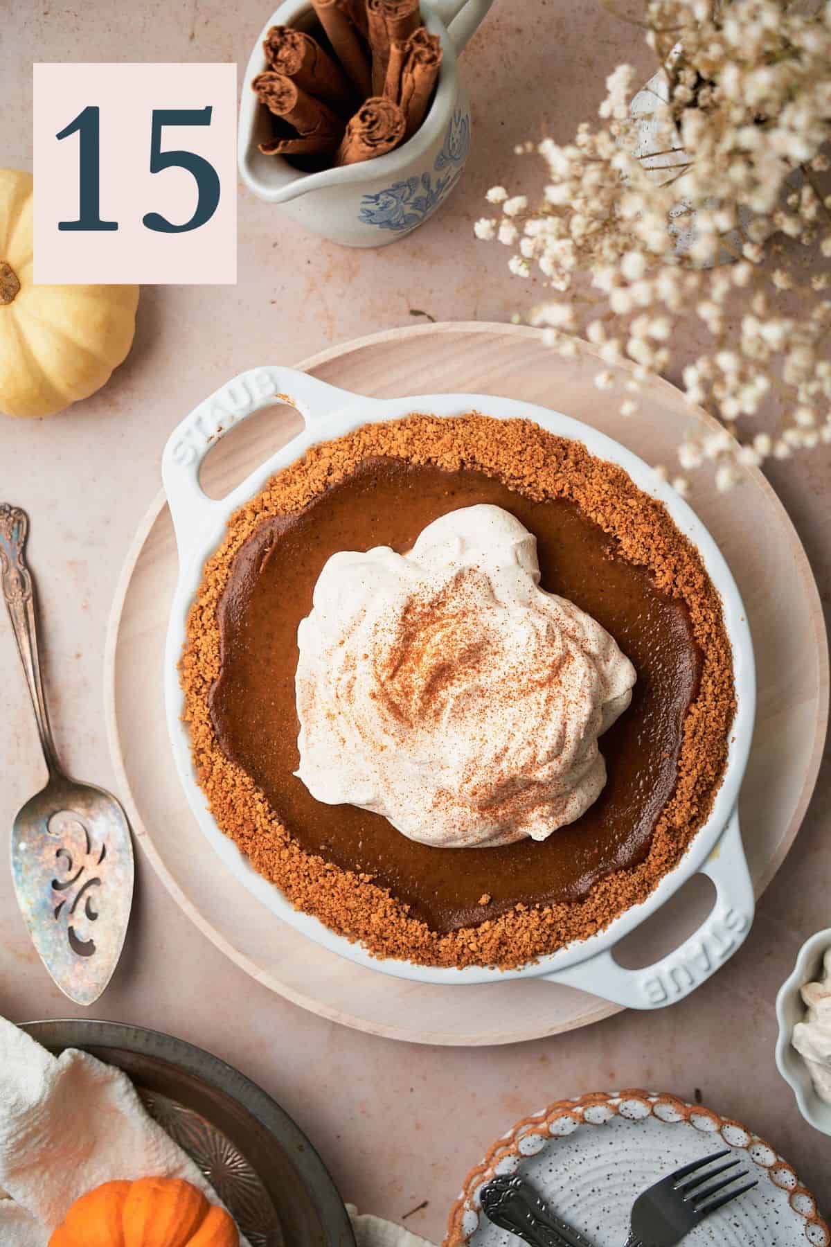 pumpkin pie topped with cinnamon maple whipped cream, and surrounded by flowers, pumpkins, and plates. 