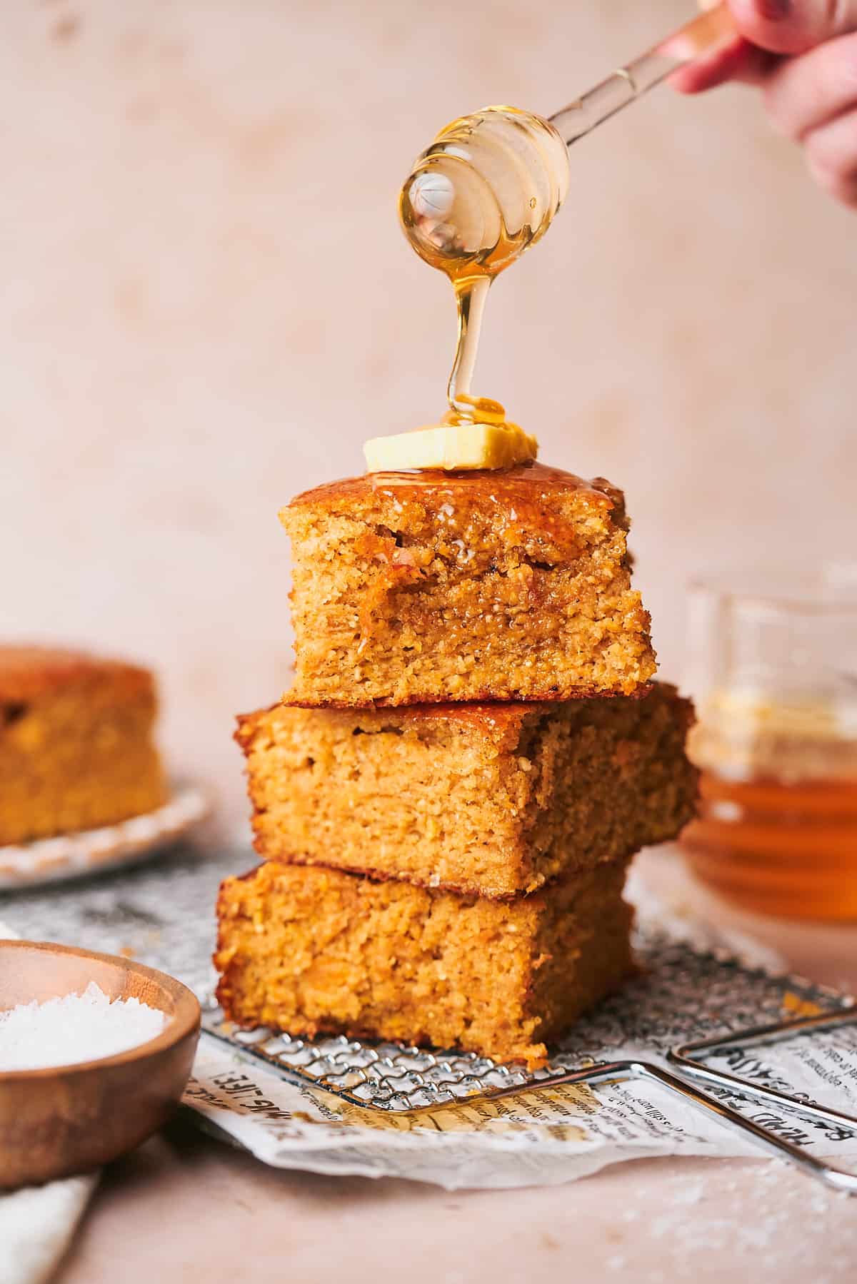 stack of sweet potato cornbread with a dollop of honey on top, and a hand drizzling honey from a honey dripper onto a stack of cornbread. 