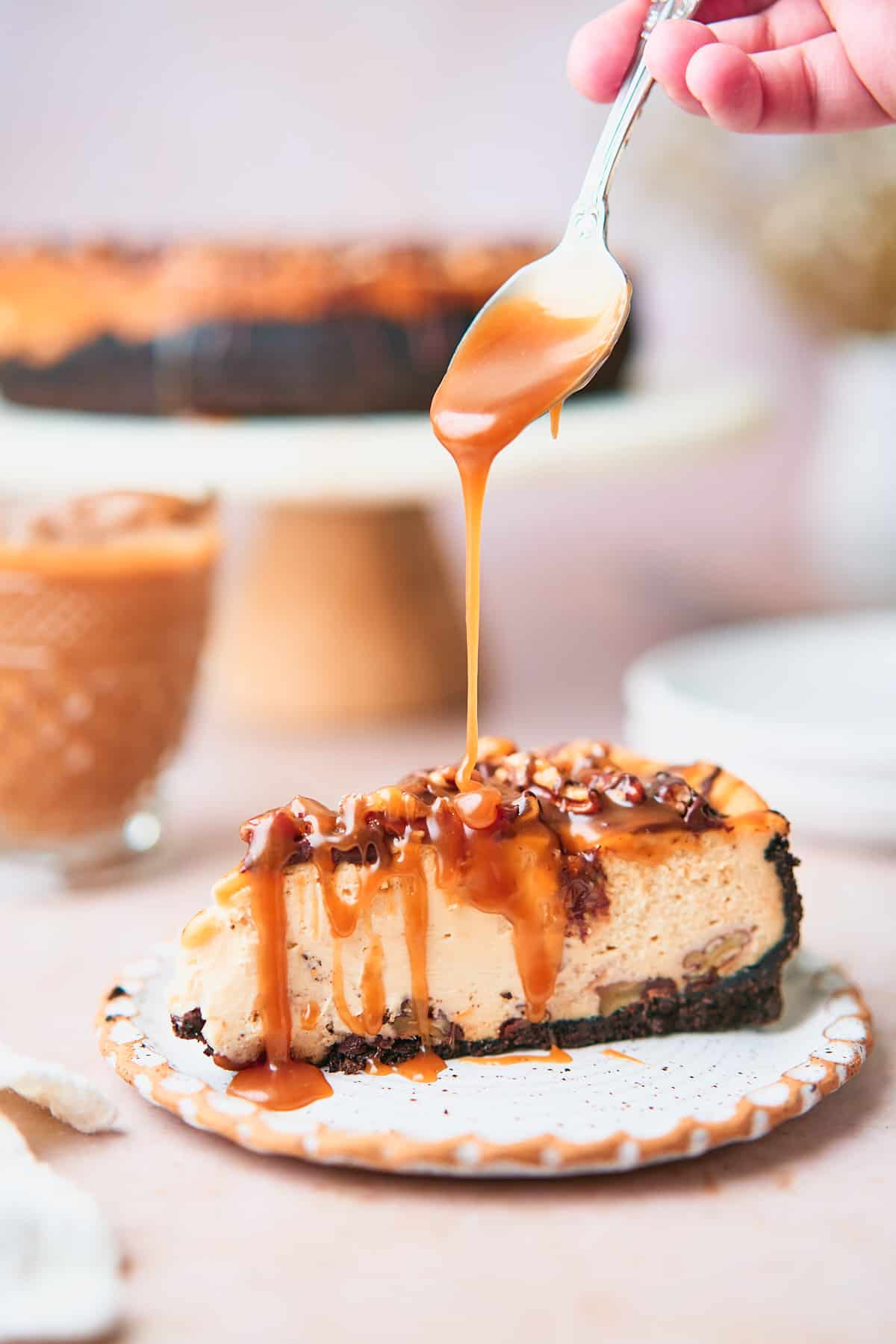 luscious slice of turtle cheesecake on a plate, with the full cheesecake in the background and a hand drizzling caramel sauce onto the cheesecake. 