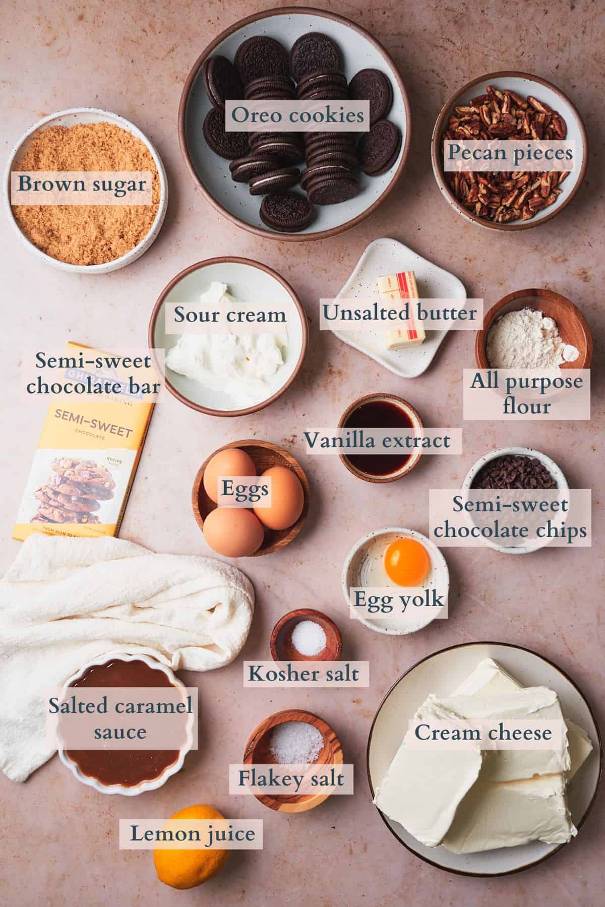 Ingredients to make turtle cheesecake laid out on a table in small bowls labeled to denote each ingredient. 