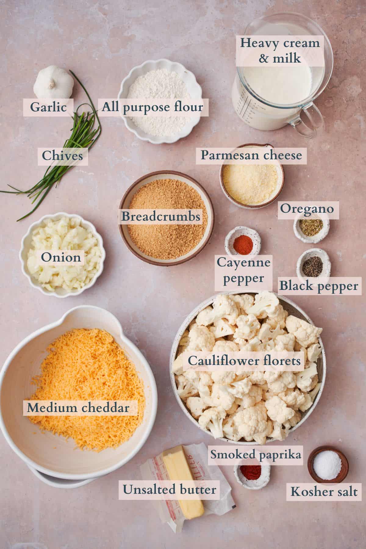 ingredients to make a cheesy cauliflower bake laid out in small bowls on a table and labeled to denote each ingredient. 