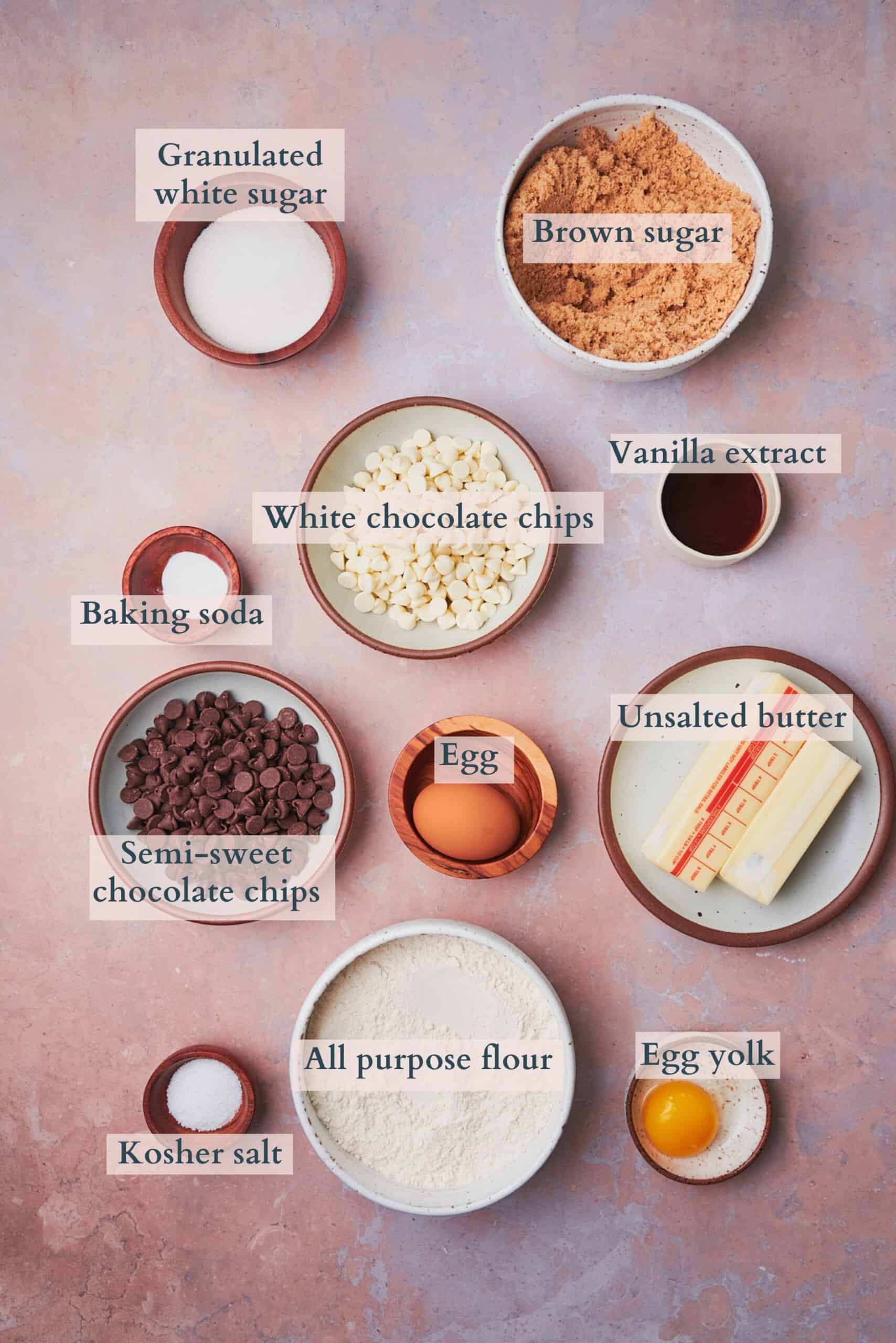Ingredients to make chocolate chip and white chocolate chip cookies laid out in small bowls and labeled to denote each ingredient.