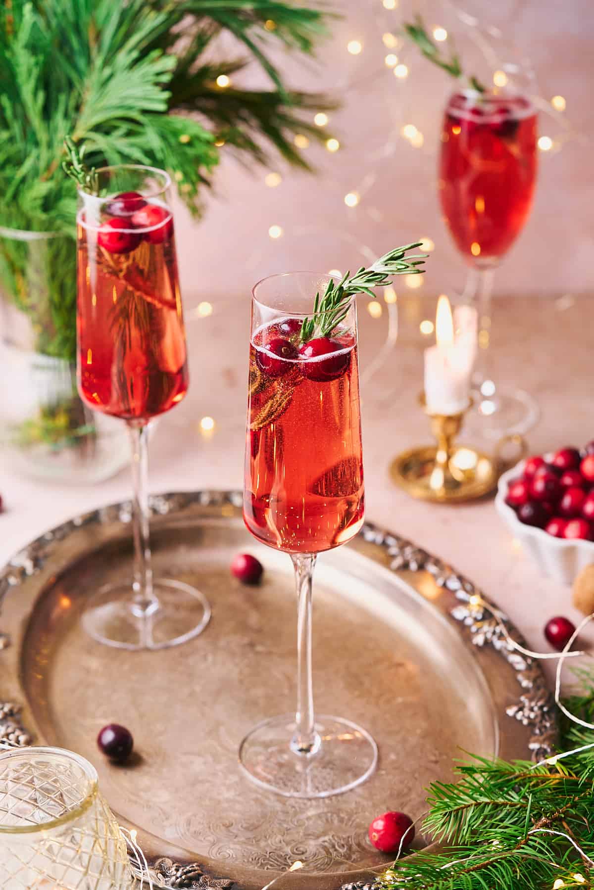 3 stunning bright red cocktails in champagne flutes with rosemary and cranberries, with garland and twinkling lights in the background.  