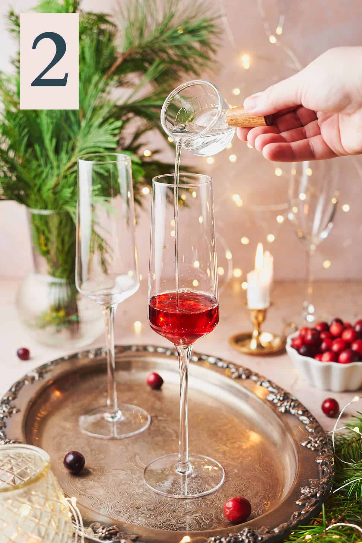 hand pouring liqueur into a champagne flute with a festive Christmas background with twinkling lights, garland, and cranberries. 
