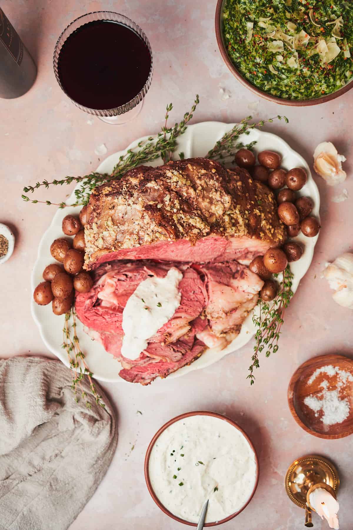 overhead view of a slow cooker prime rib on a serving platter with fresh thyme, and drizzled with creamy horseradish sauce, surrounded by wine, potatoes, creamed spinach, and more. 