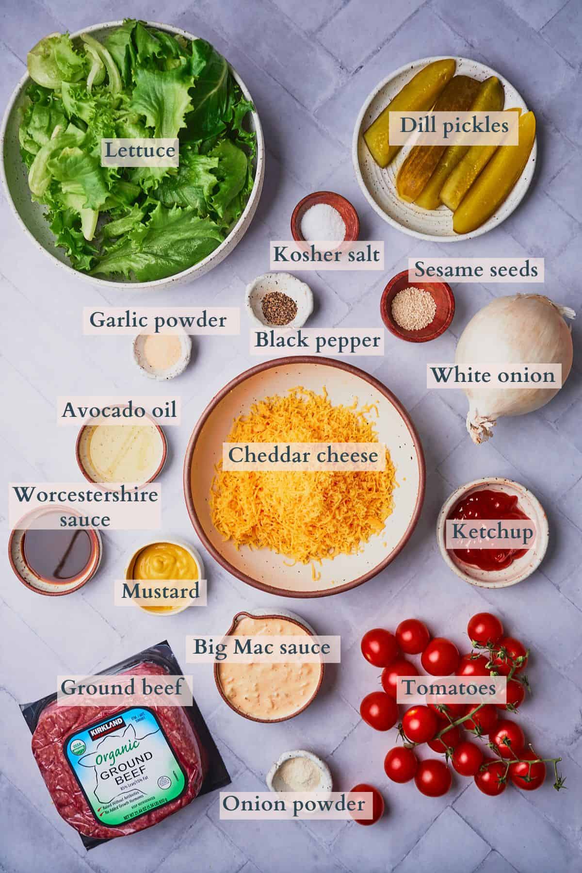 Ingredients to make a big Mac salad laid out on a table in small bowls and on plates and labeled to denote each ingredient.