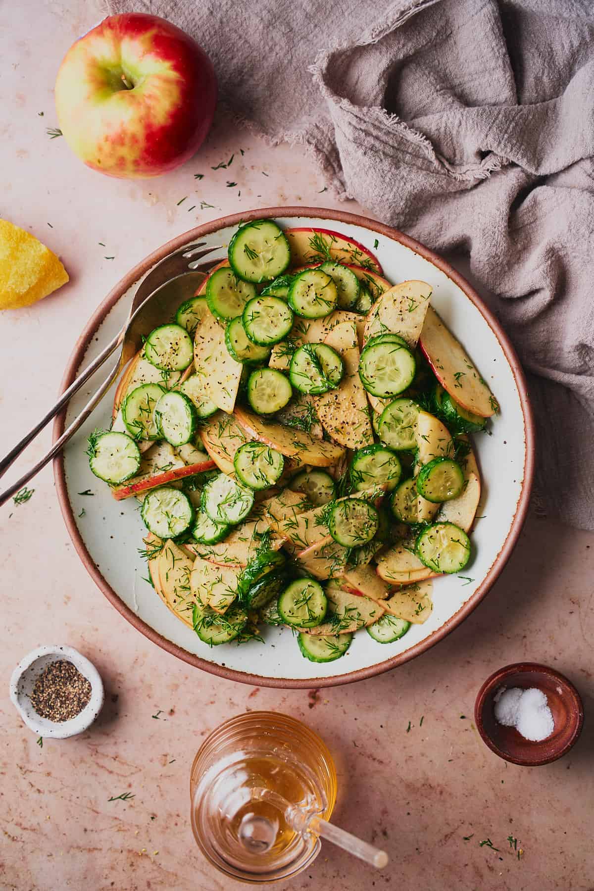 Apple and cucumber salad on a rounded serving plate, surrounded by honey, fresh apples, lemon, salt, and pepper. 