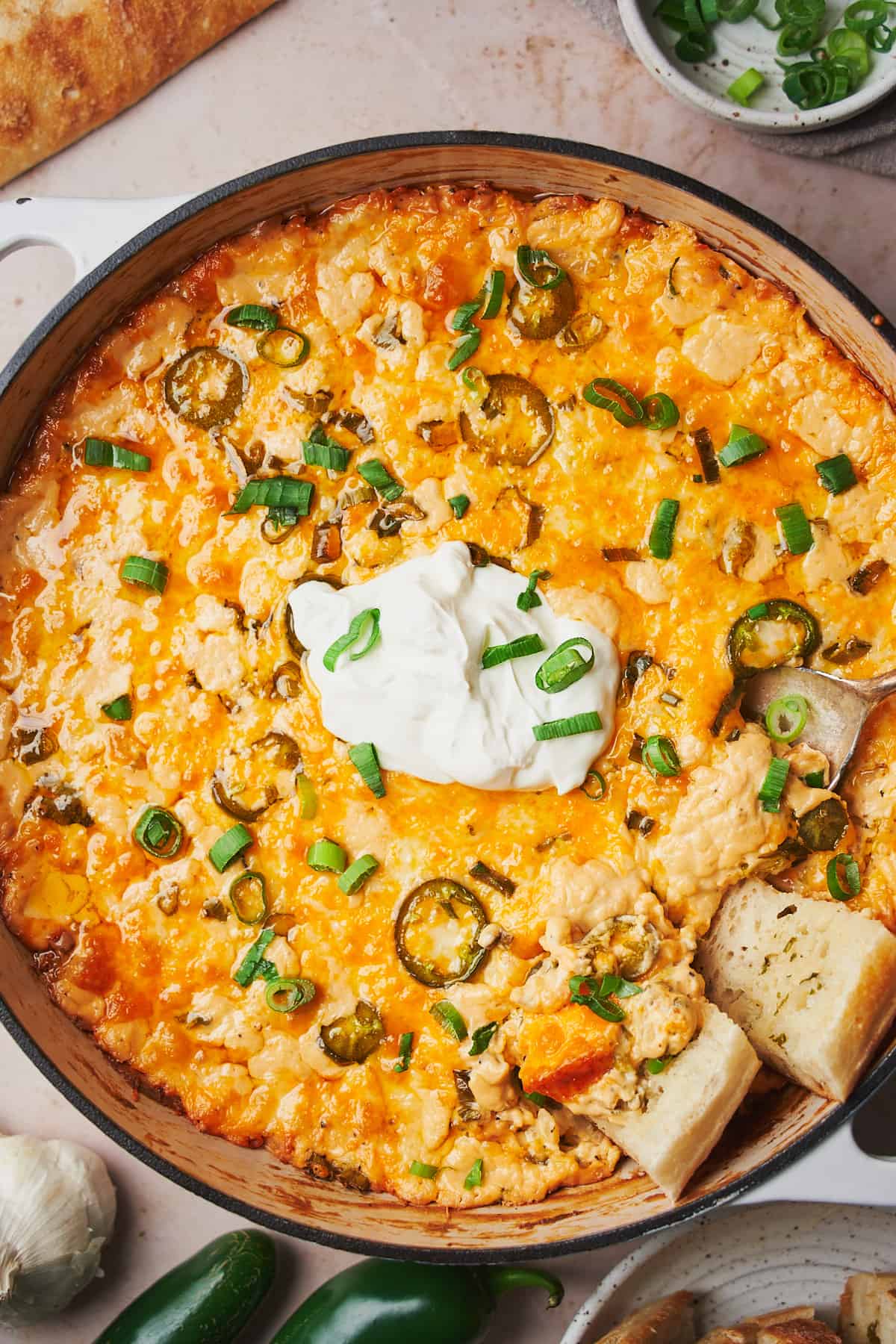 baked jalapeno dip in an enameled cast iron skillet topped with sour cream and green onions in the center, with grilled bread dipped in the dip. 