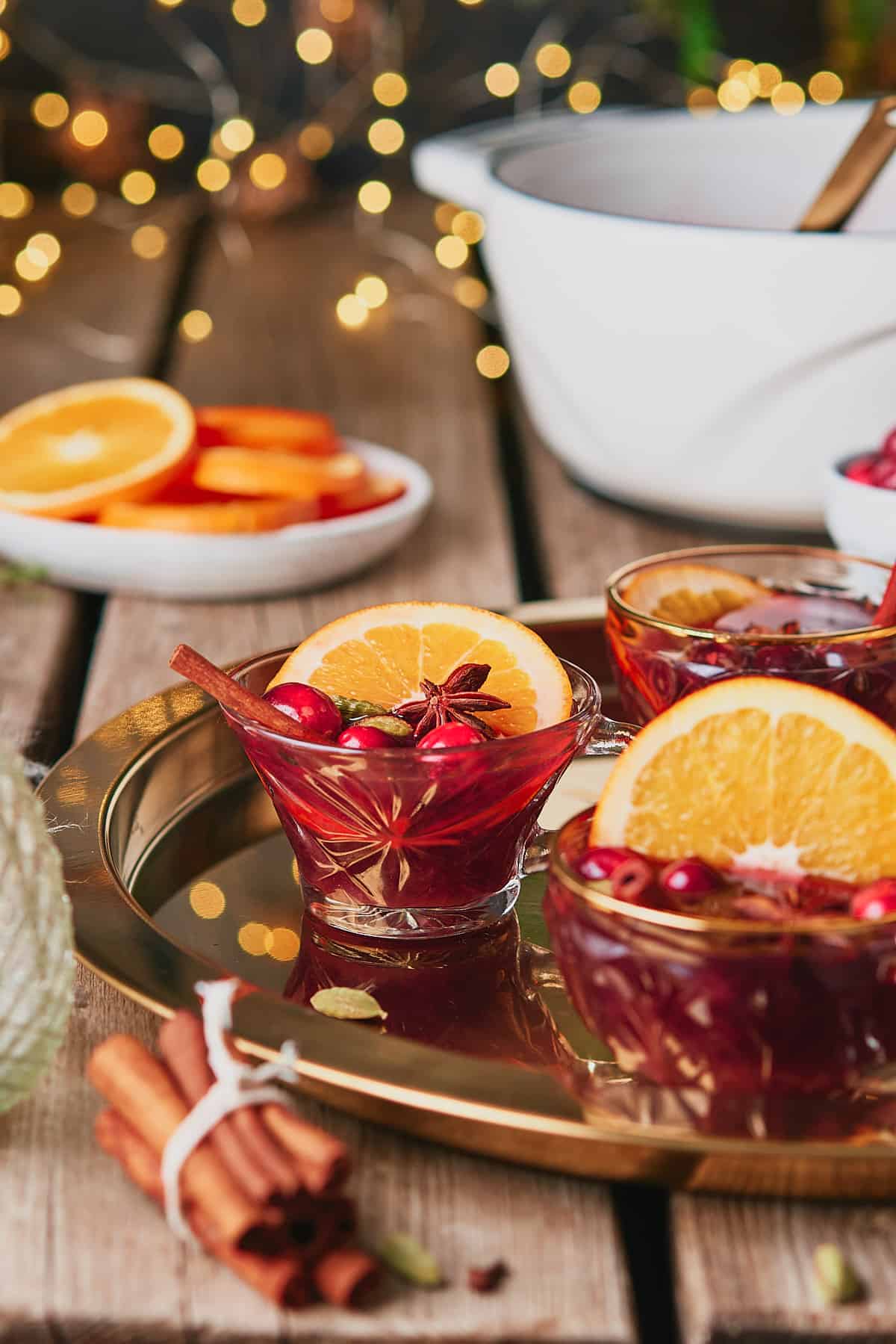 Glasses of mulled red wine and gin on a gold platter, on a wooden table with twinkly lights, oranges and a stock pot in the background. 