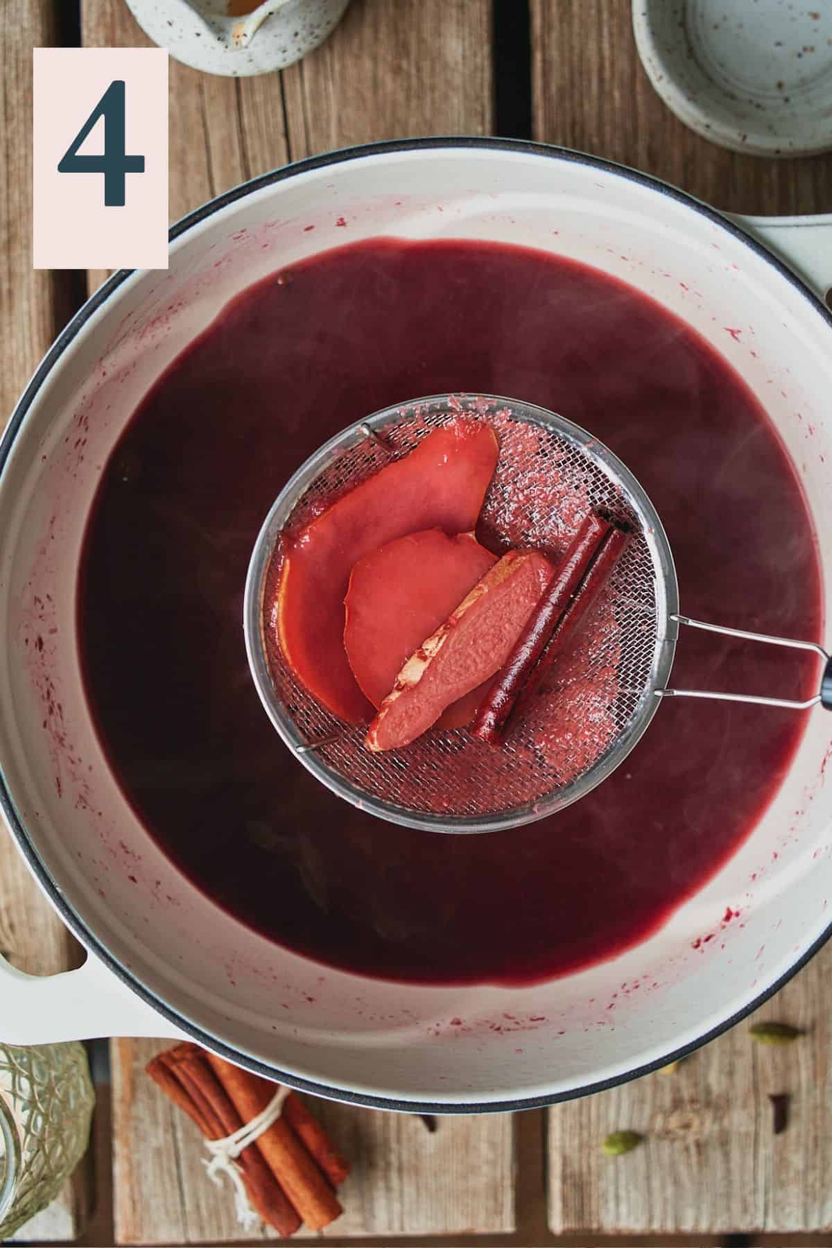 using a small skimmer to strain out the fruit and whole spices from a mulled wine mixture in a Dutch oven. 