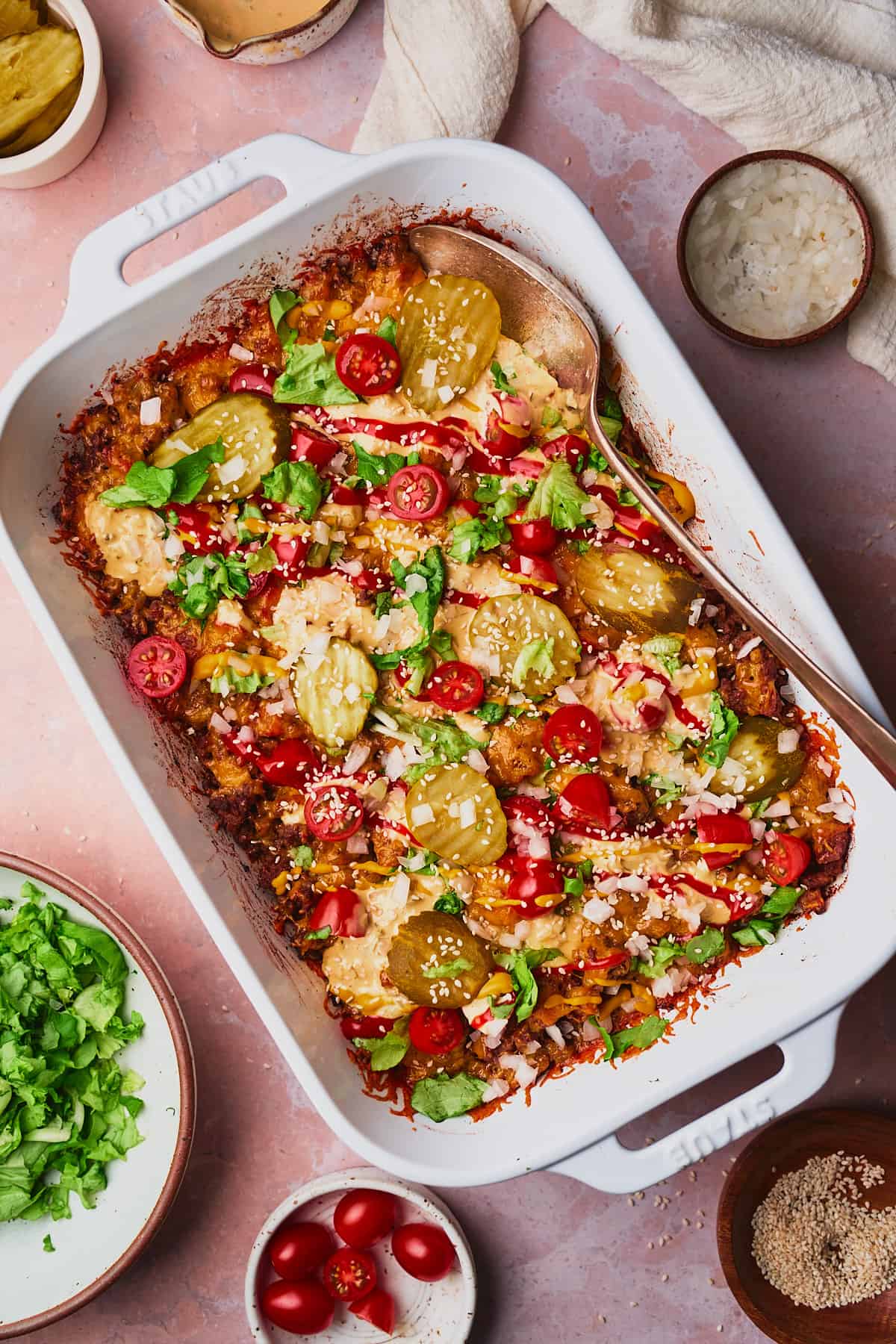 Flat lay shot of a big mac casserole topped with pickles, lettuce, tomatoes, onions, and more surrounded by the ingredients used to make the casserole. 