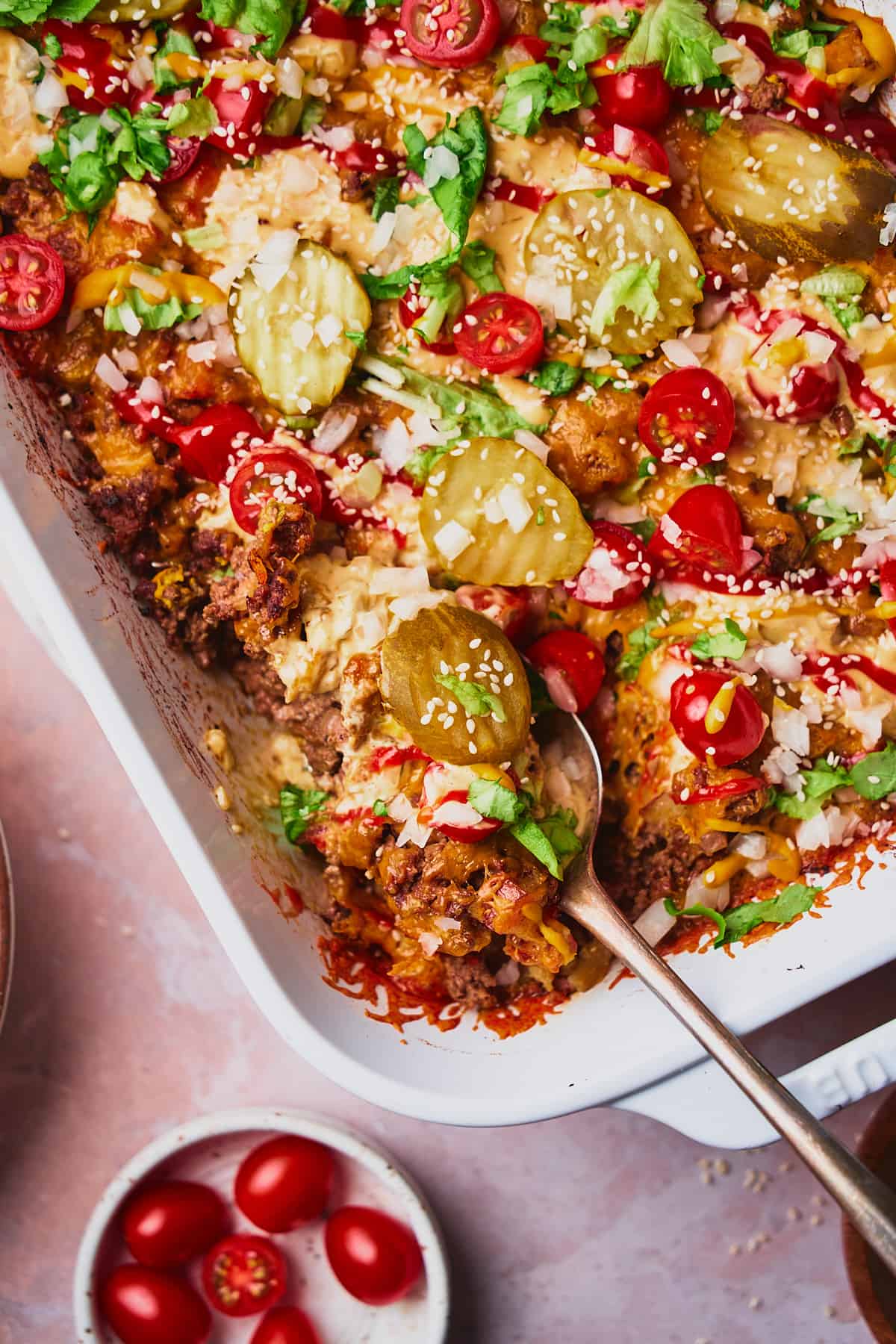 Close up shot of big mac casserole in the corner of a white rectangular casserole dish topped with pickles, tomatoes, lettuce, ketchup and mustard. 