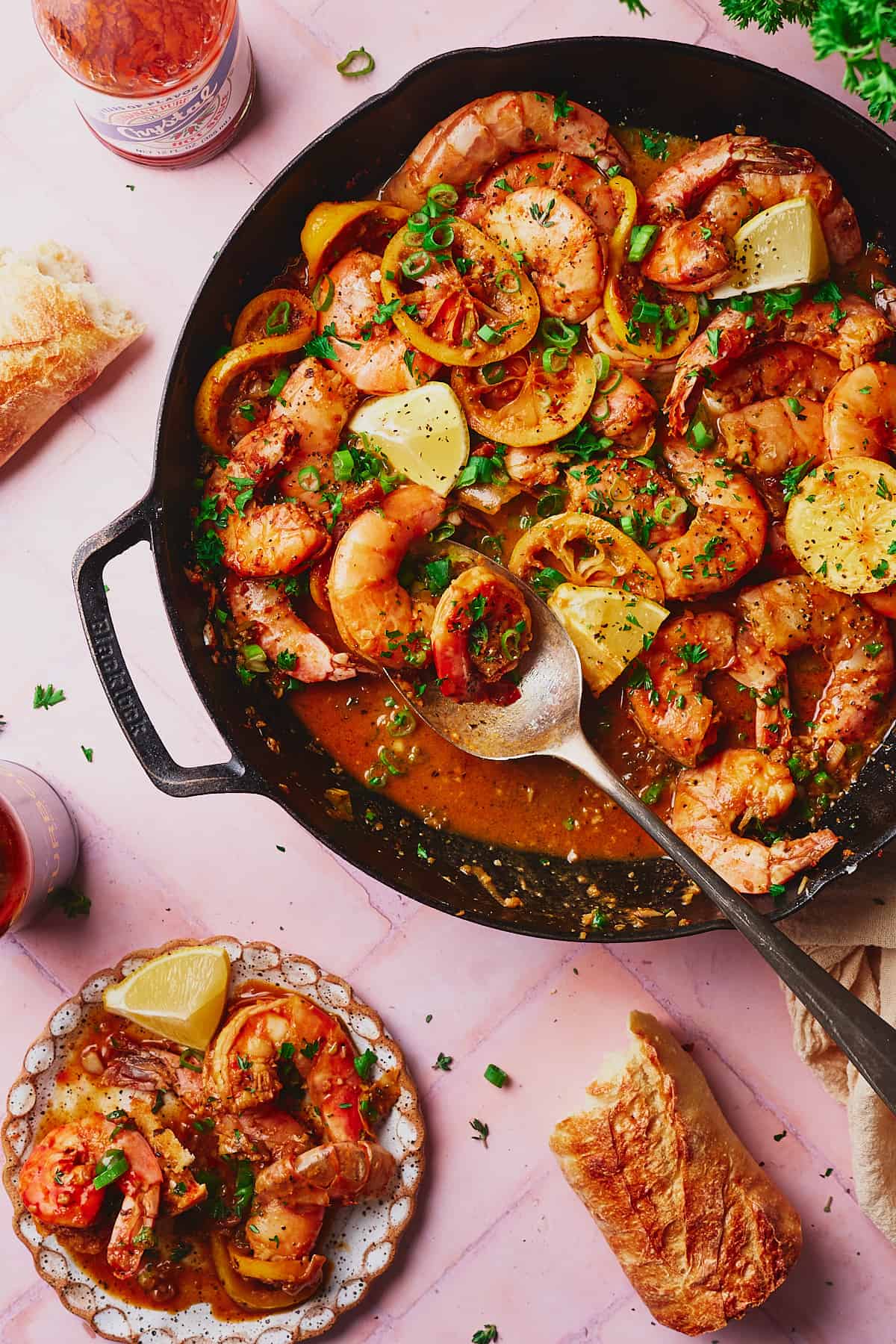 New Orleans BBQ Shrimp in a cast iron skillet, with some on a plate with fresh lemons, a baguette, hot sauce, beer, and parsley on the scene. 