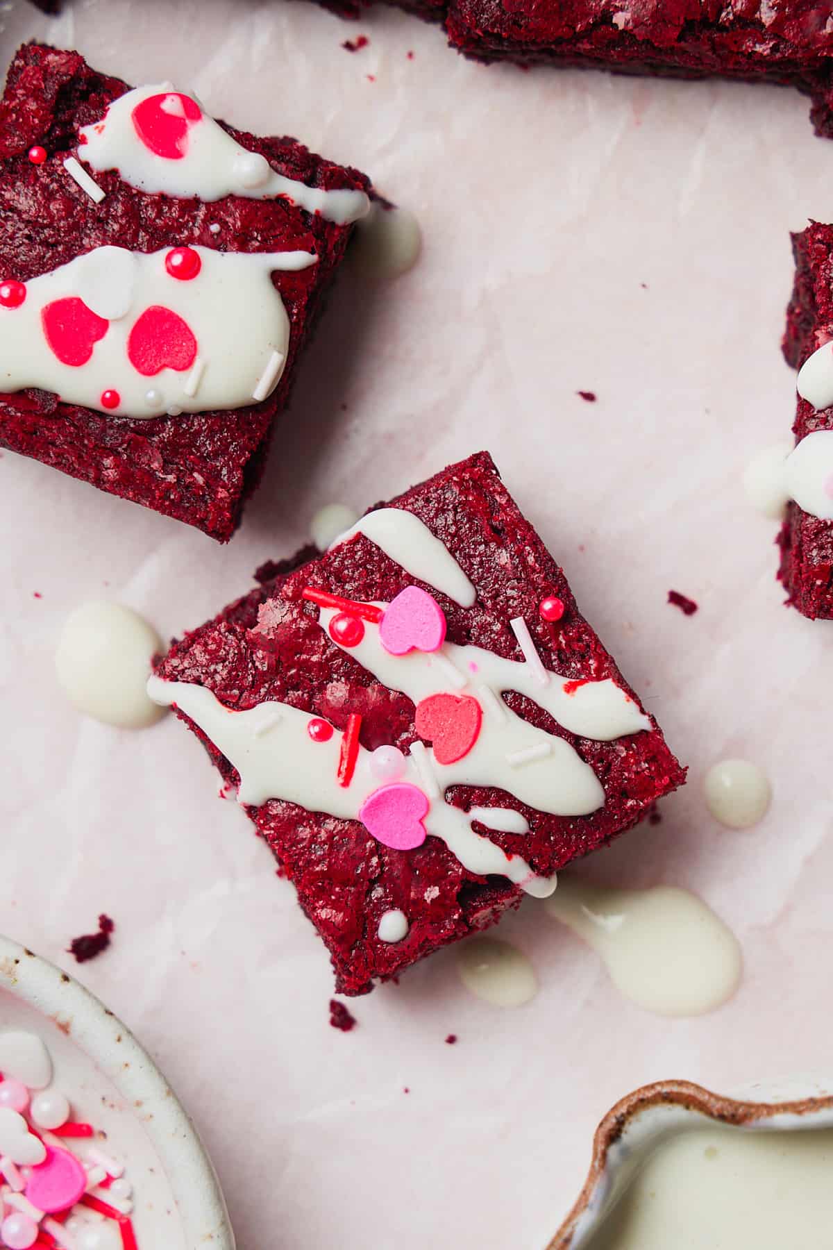 Flat lay photo of a red velvet brownie drizzled with cream cheese frosting with Valentine's Day sprinkles surrounded by other brownies on white parchment. 