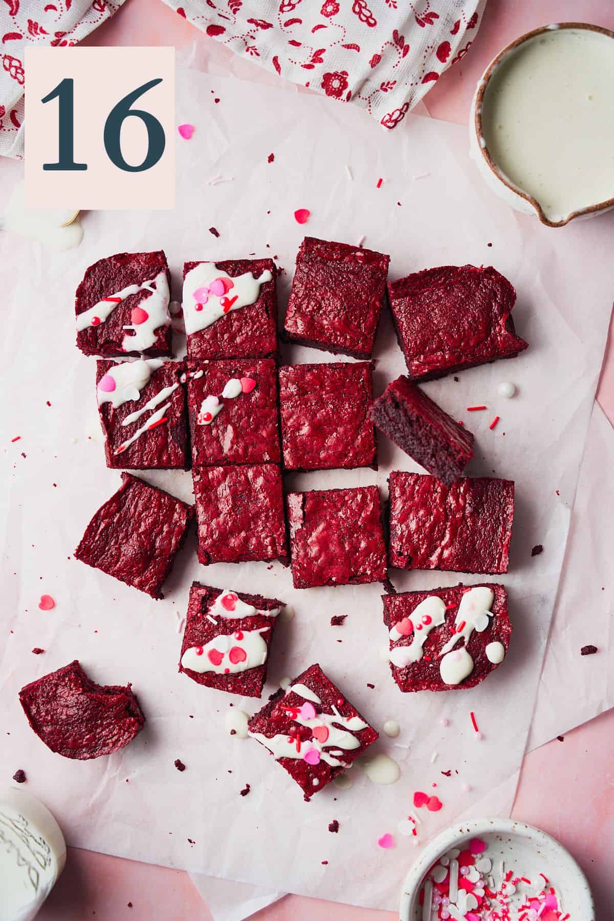 Red velvet brownies on white parchment on a pink backdrop drizzled with cream cheese frosting and sprinkles. 