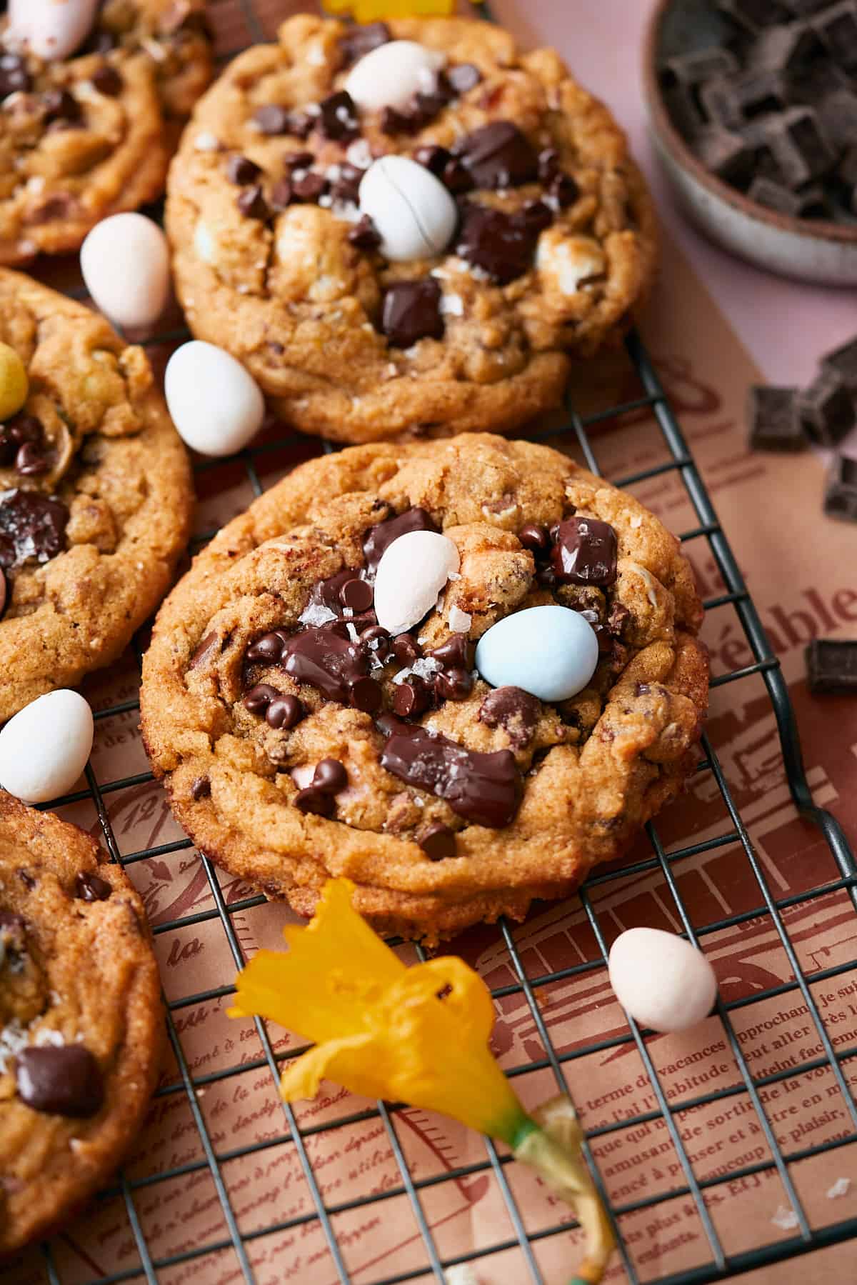 Close up shot of a Cadbury egg chocolate chip cookie on a cooling rack surrounded by extra mini Cadbury eggs, cookies and spring daffodils. 