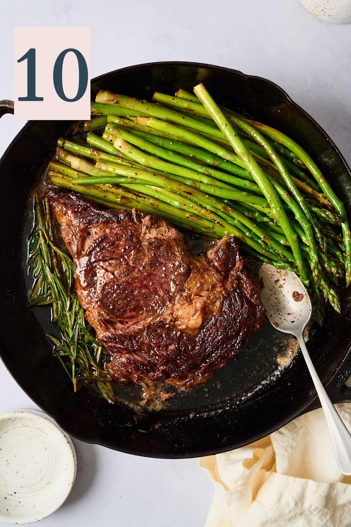 Steak in a skillet with rosemary, a spoon and asparagus added.