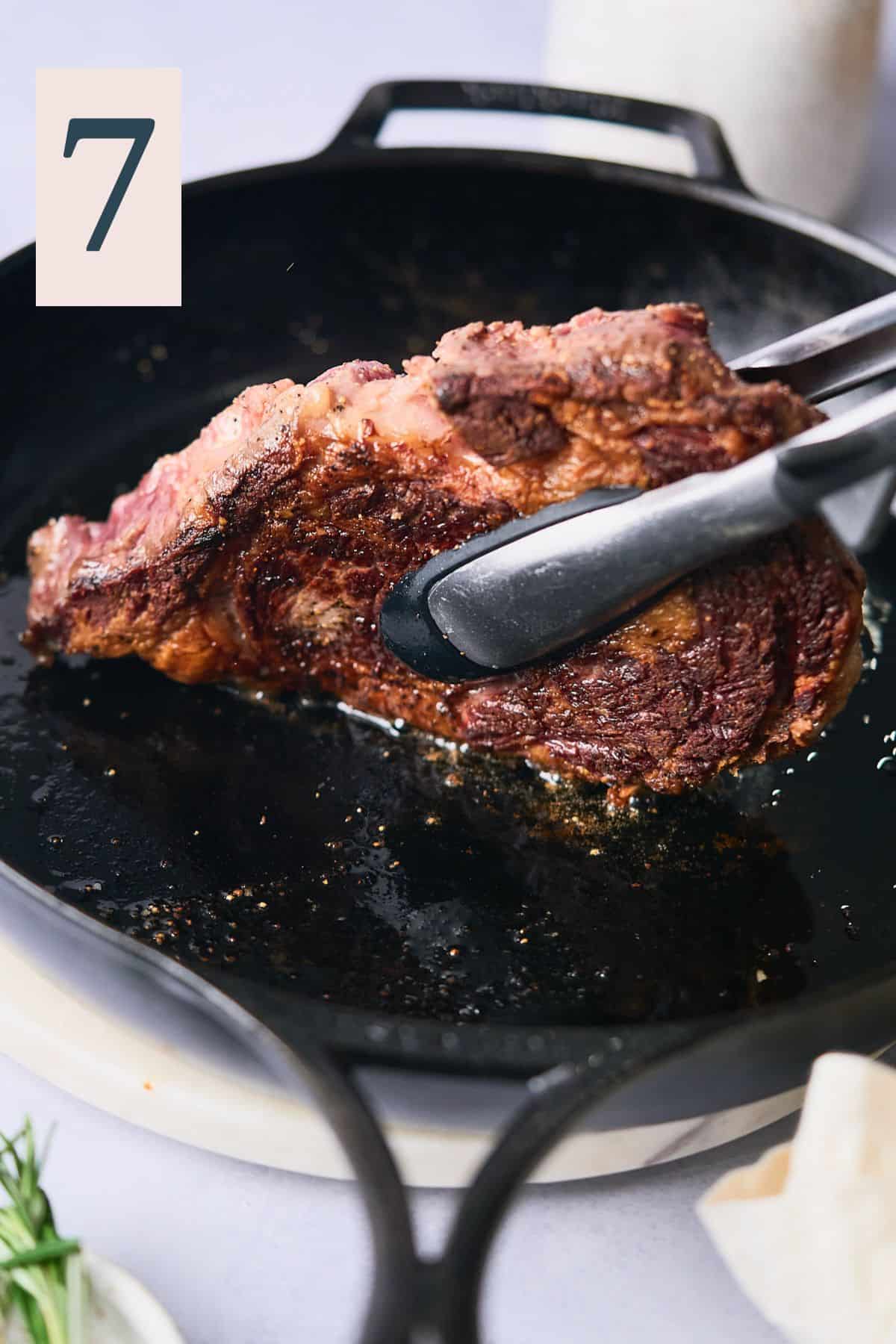 Tongs holding up a ribeye steak on one side to cook the fat cap and other uncooked parts of the side of the steak. 