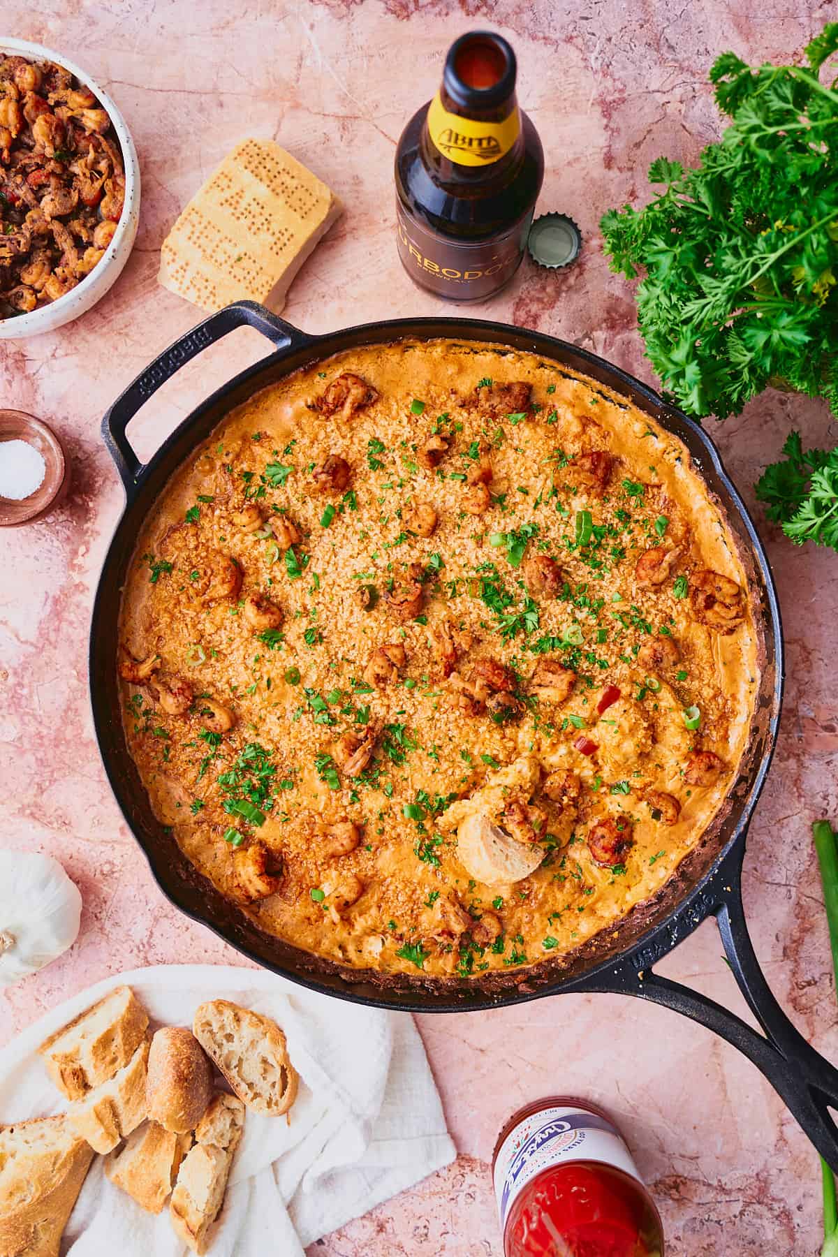 Baked hot crawfish dip in a cast iron skillet and topped with more cooked crawfish, parsley, green onions, and surrounded by bread, hot sauce, beer, and more.