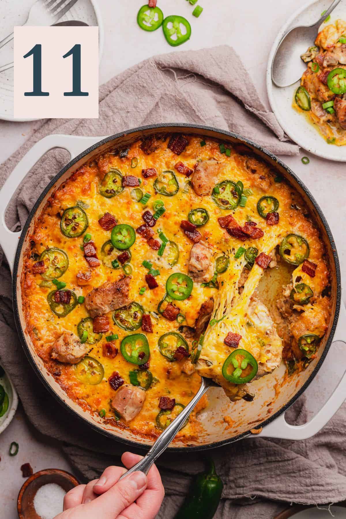 hand pulling cheesy jalapeno popper chicken from a skillet surrounded by plates with casserole on them and ingredients to make the dish. 