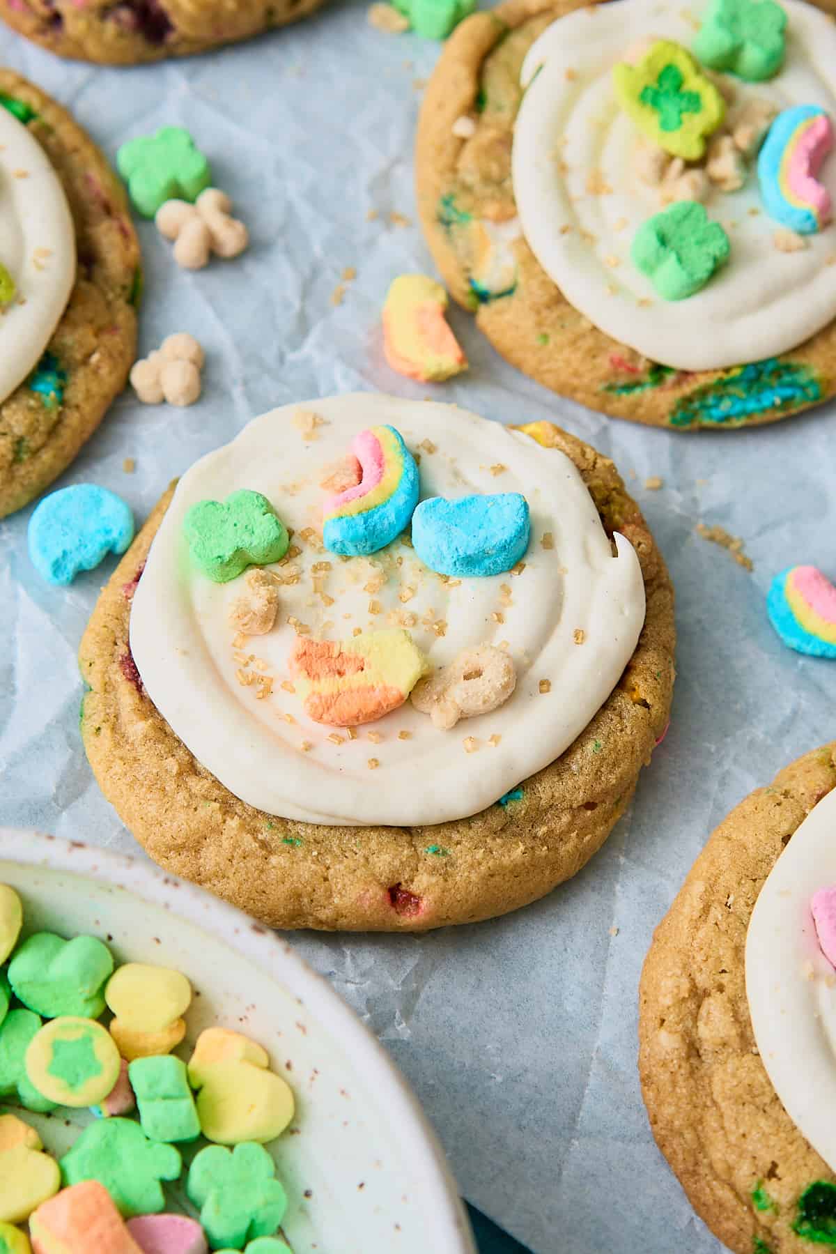 Close up shot of Lucky Charms cookies with frosting and topped with cereal and gold sprinkles on a parchment paper sheet surrounded by the other cookies and cereal. 